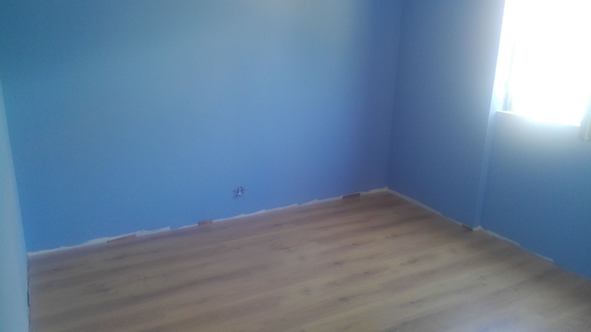 Quarto vazio com paredes azuis e piso de madeira, com uma janela à direita.