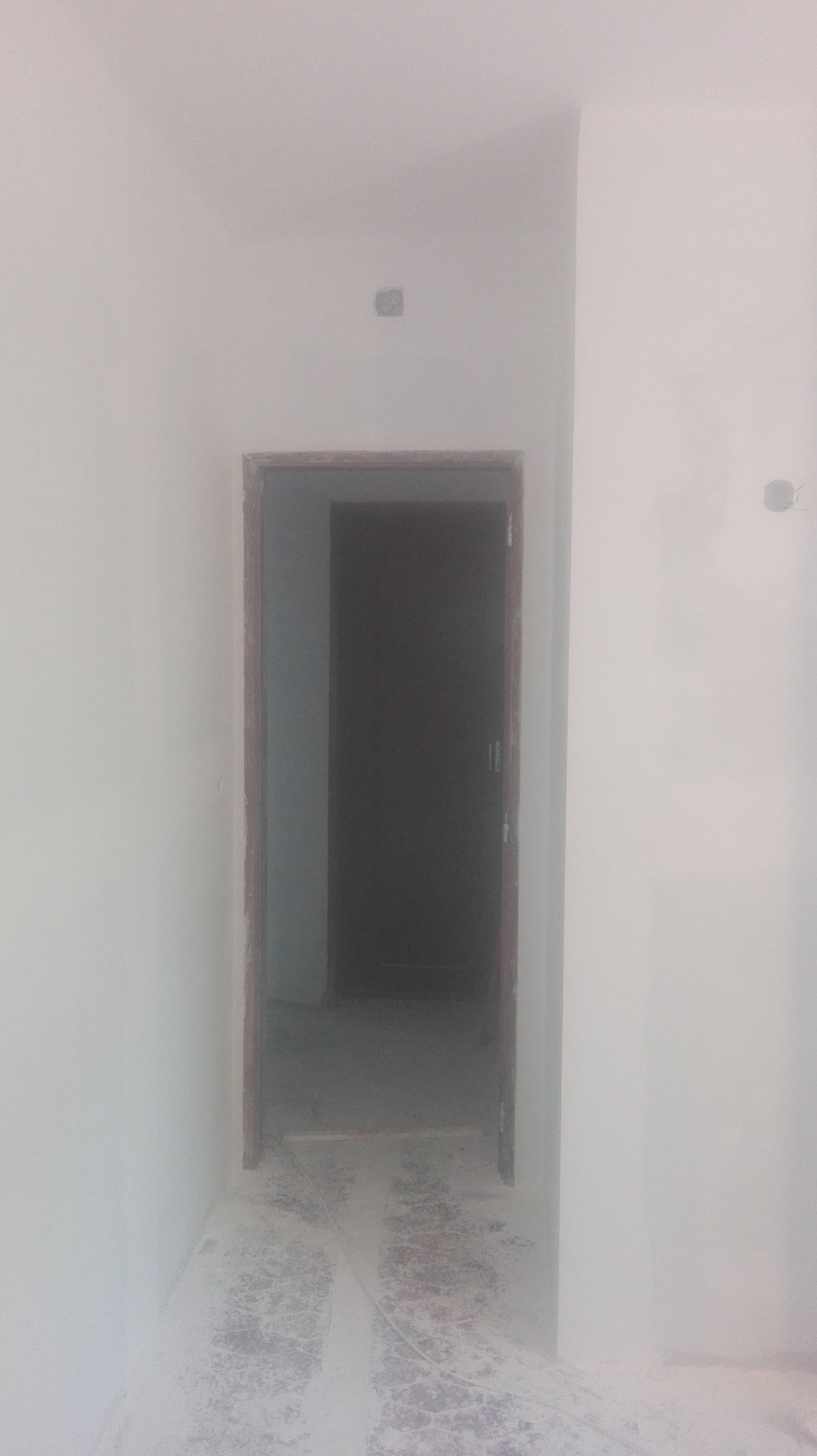 Corredor estreito com uma porta no final, que dá acesso a um quarto escuro. Paredes e chão brancos.