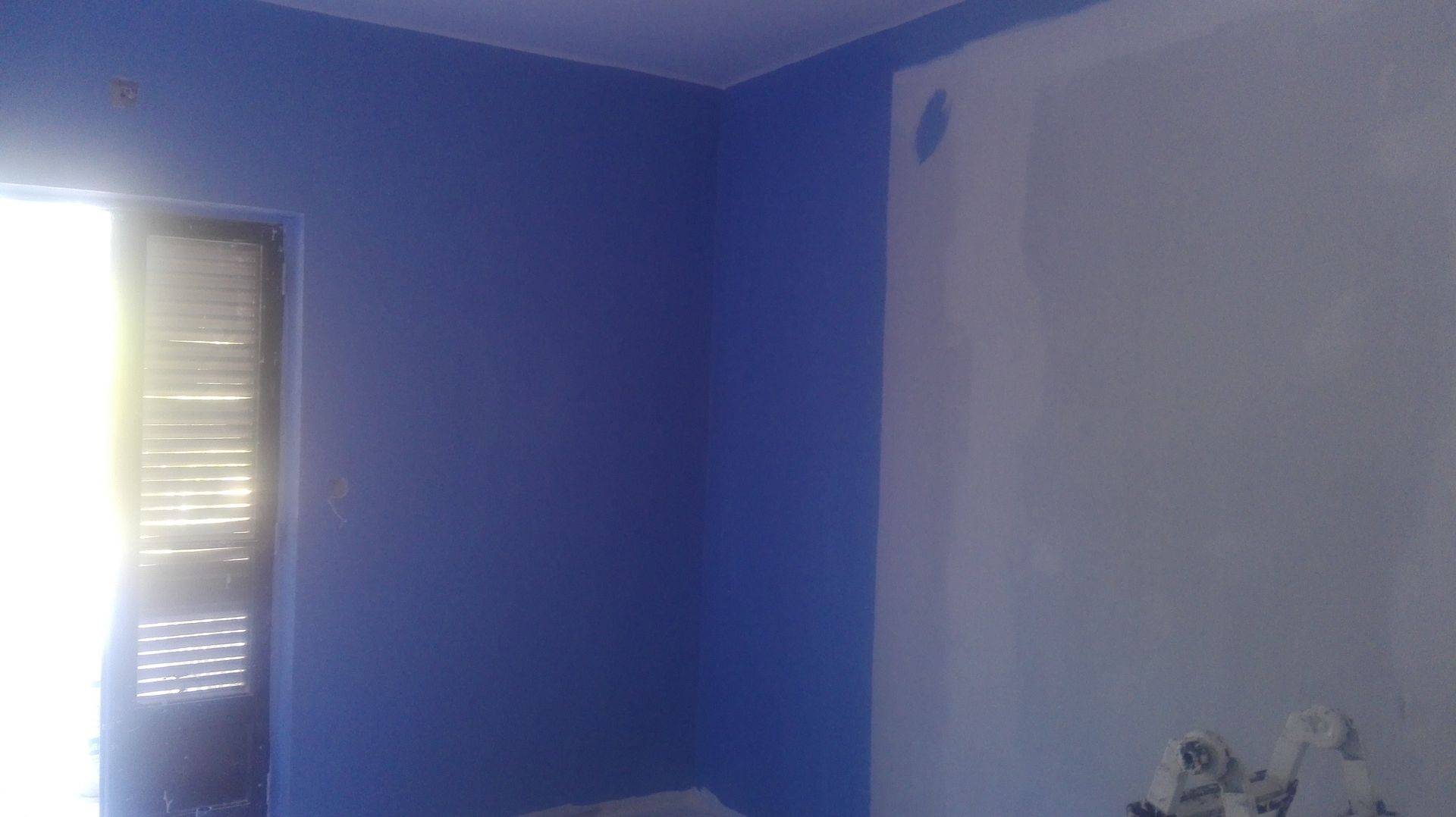 Quarto de canto, uma parede pintada de azul, a outra parede branca sem acabamento; porta com persianas.