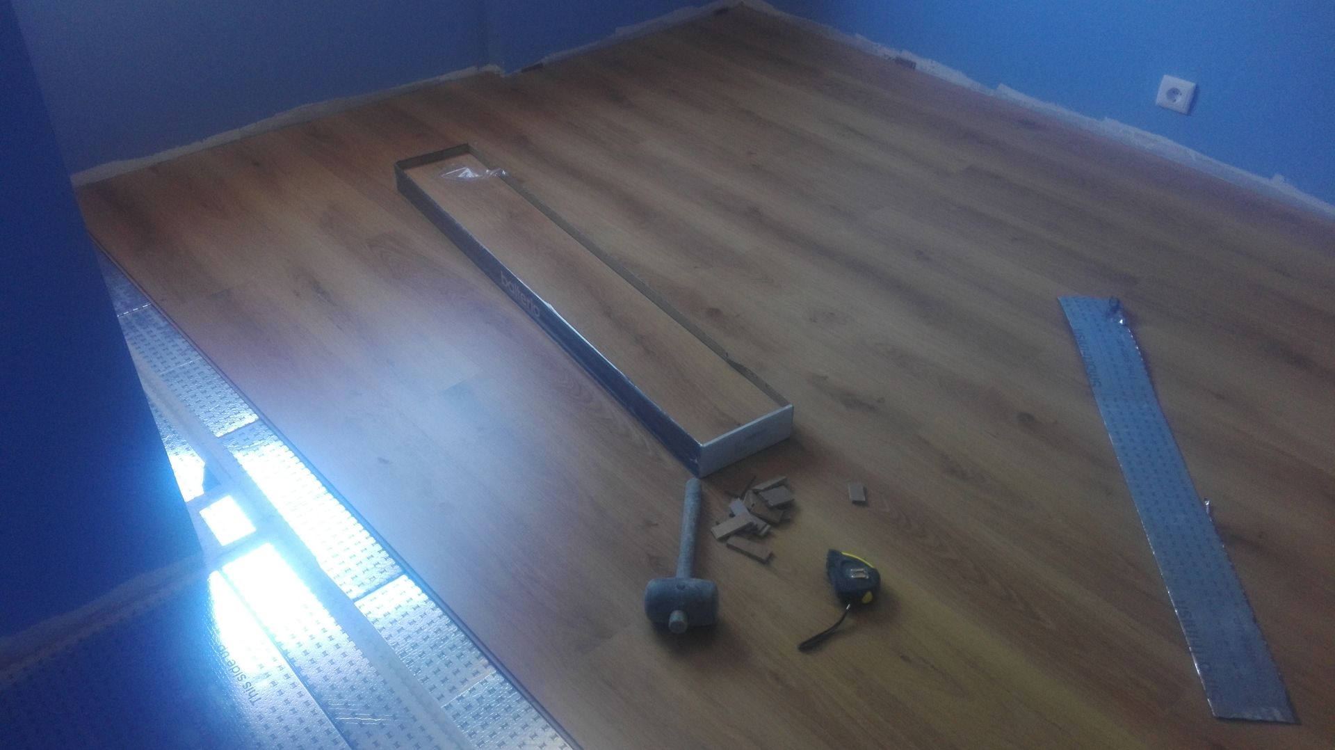 Instalação de piso laminado em andamento; ferramentas e materiais sobre um piso parcialmente acabado, parede azul ao fundo.