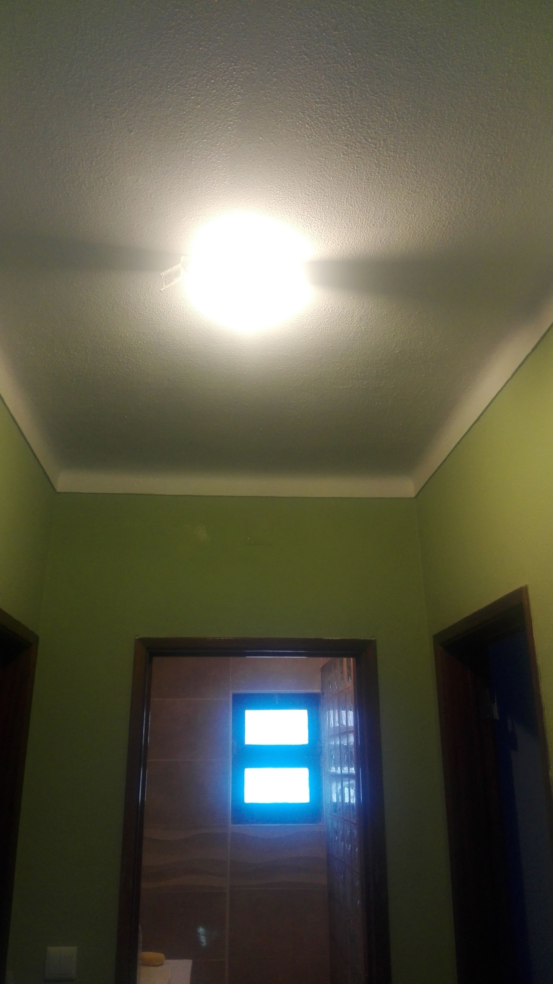 Corredor verde com uma luz forte no teto; a porta no final tem uma janela iluminada.
