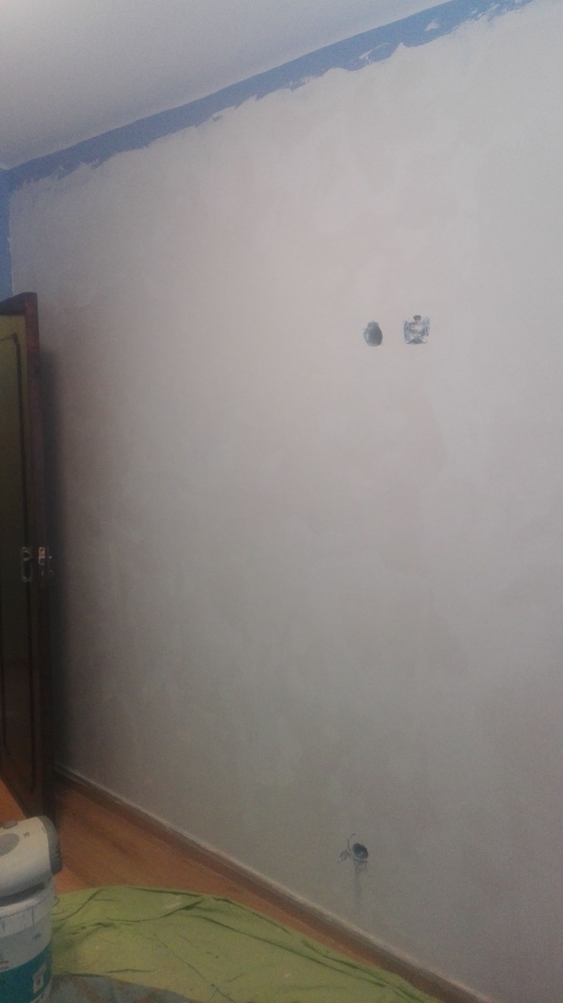 Parede branca com detalhes em azul, dois furos e uma tomada. Uma porta, o chão e uma lata de tinta são visíveis.