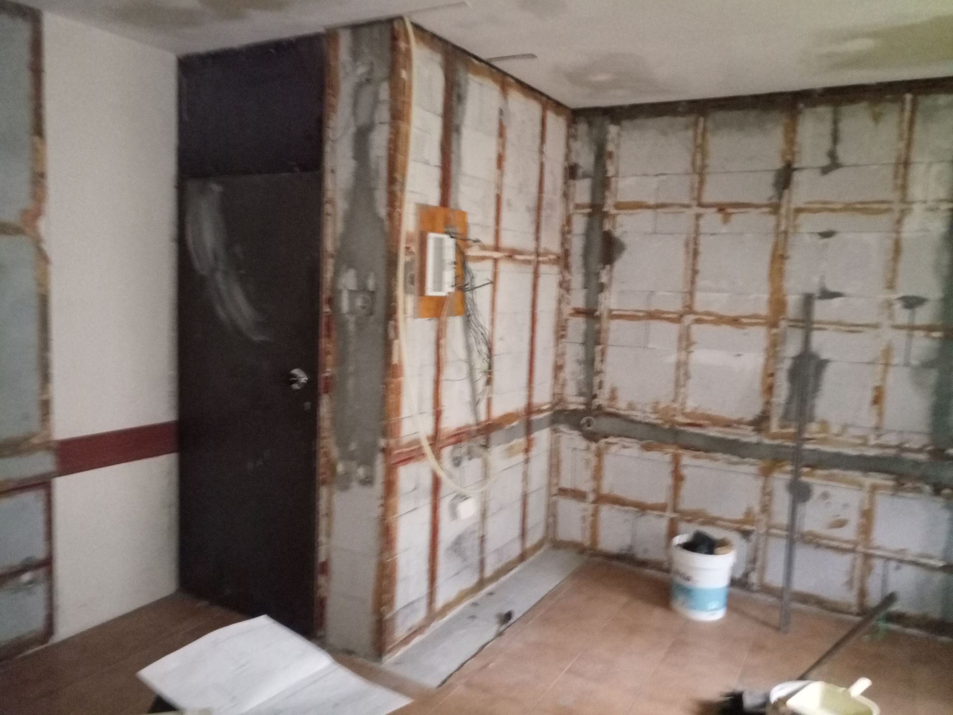 Quarto interior em construção, com vigas de sustentação expostas. Porta escura, balde branco, remendos de cimento e piso aberto.