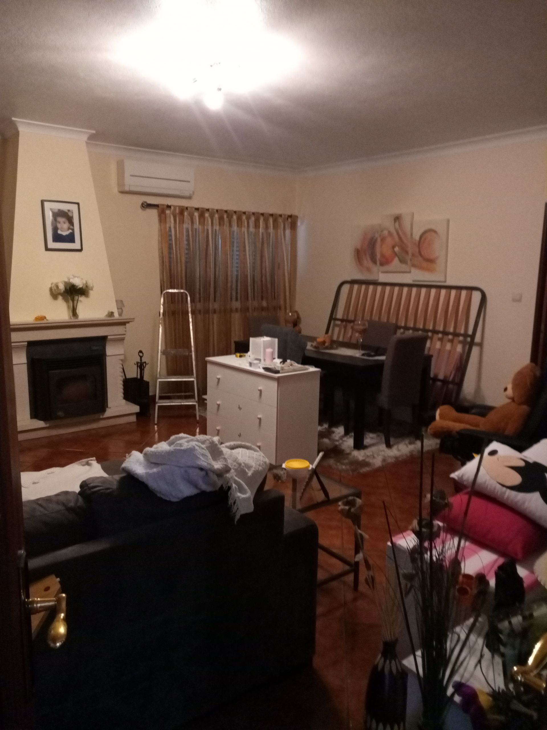 Sala de estar desarrumada com lareira, piano e estrutura de cama encostada na parede.