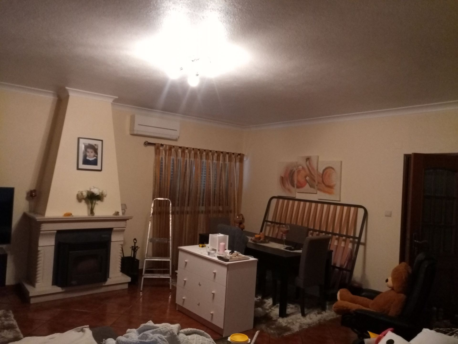 Sala de estar com lareira, cortinas, cômoda e piano. Um ursinho de pelúcia está sentado em uma cadeira.