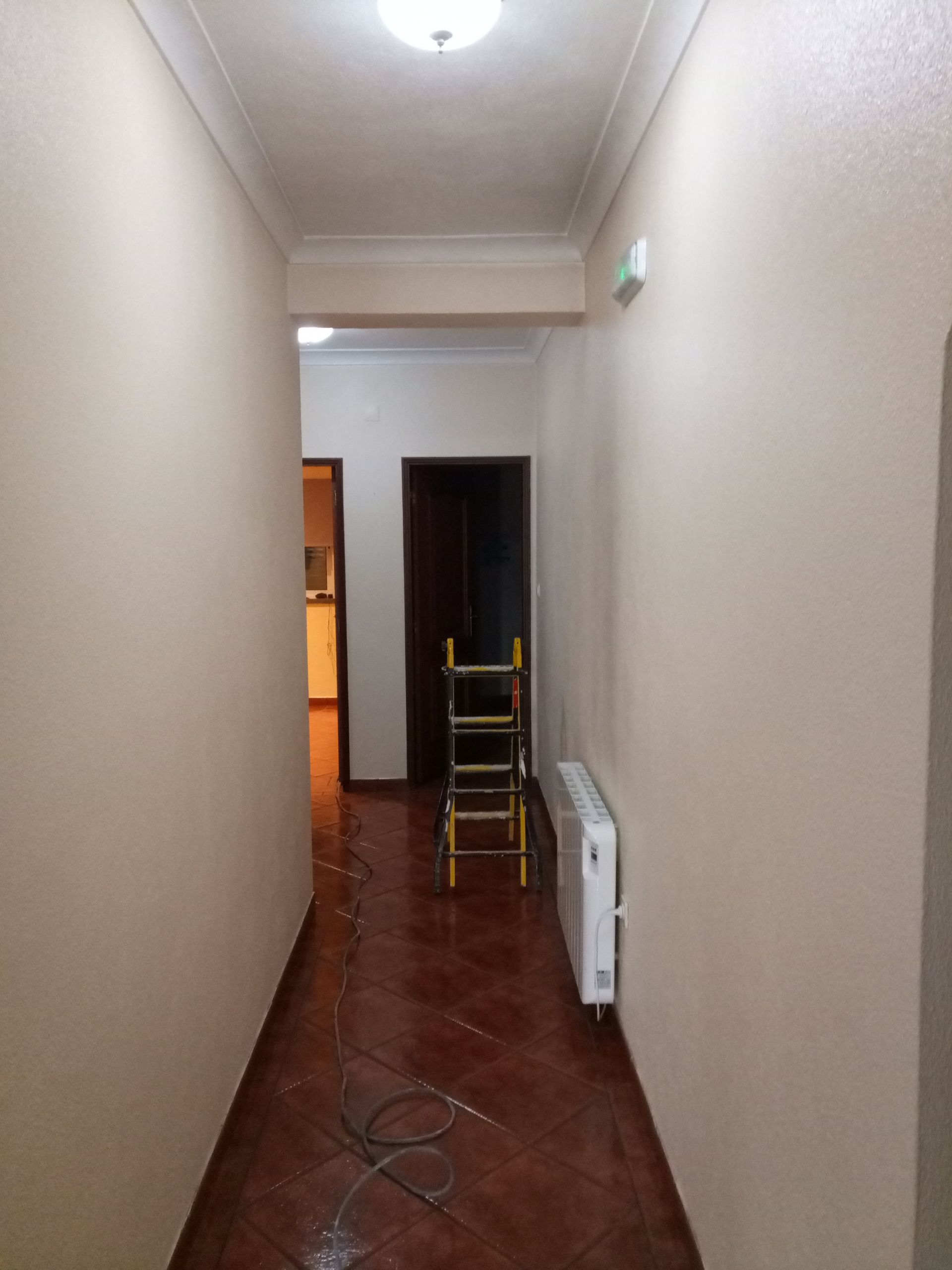 Corredor estreito com piso de ladrilho marrom, paredes bege claro e uma porta marrom fechada no final.