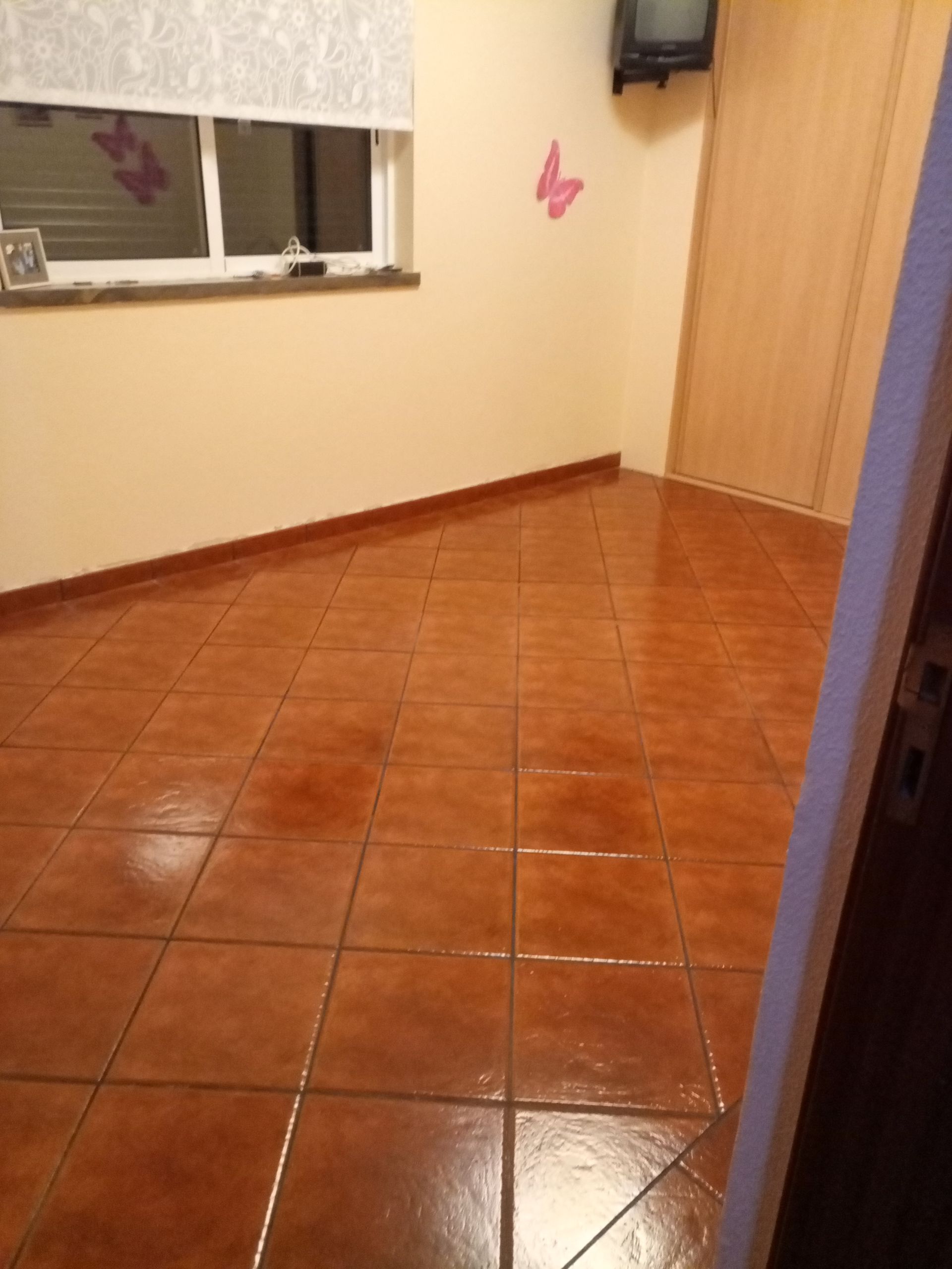 Quarto vazio com piso de azulejos laranja, paredes bege, janela pequena e armário de madeira.