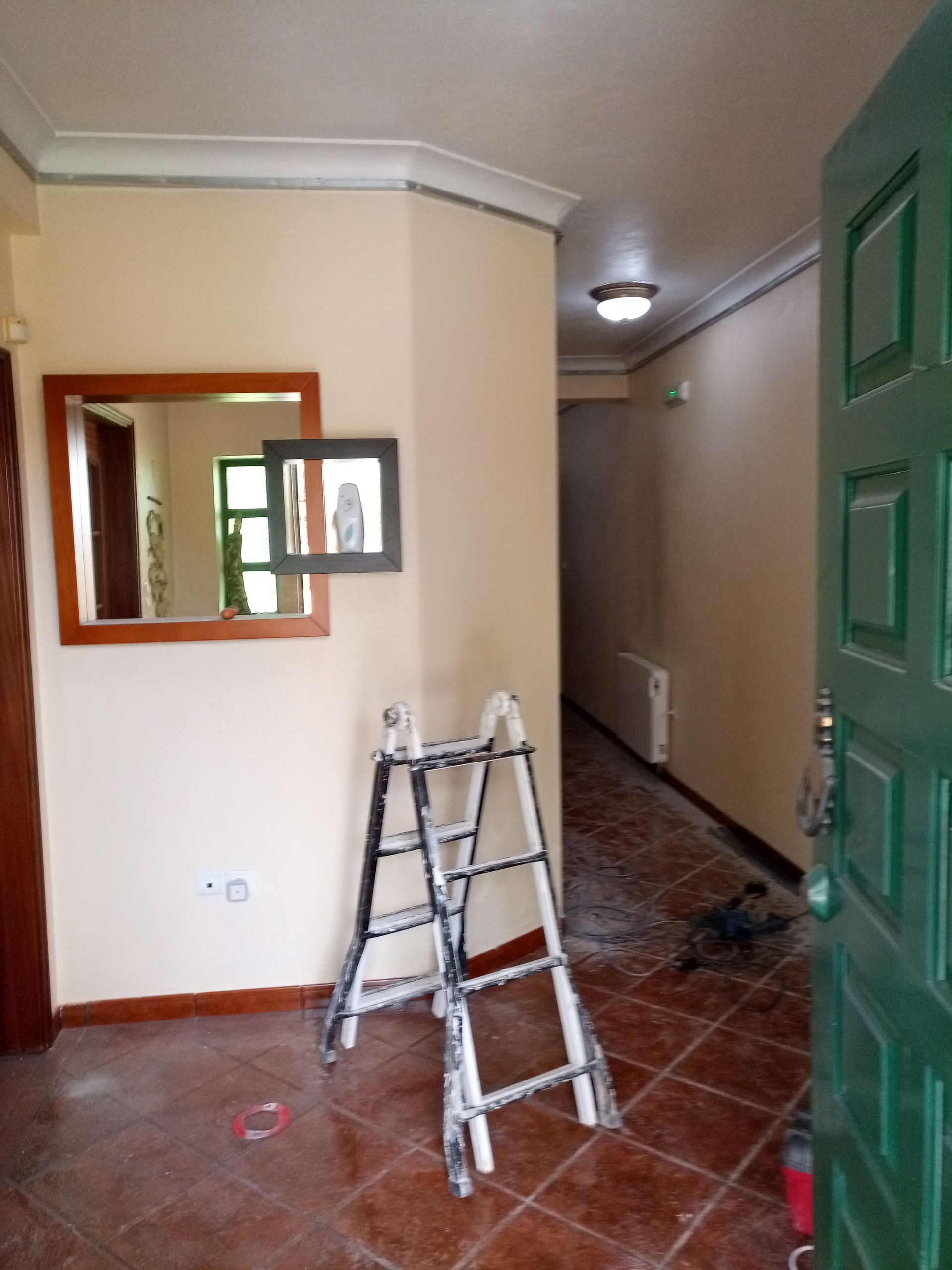 Hall de entrada com piso de azulejo marrom, paredes bege, porta verde e escada encostada na parede. Um espelho está pendurado na parede.