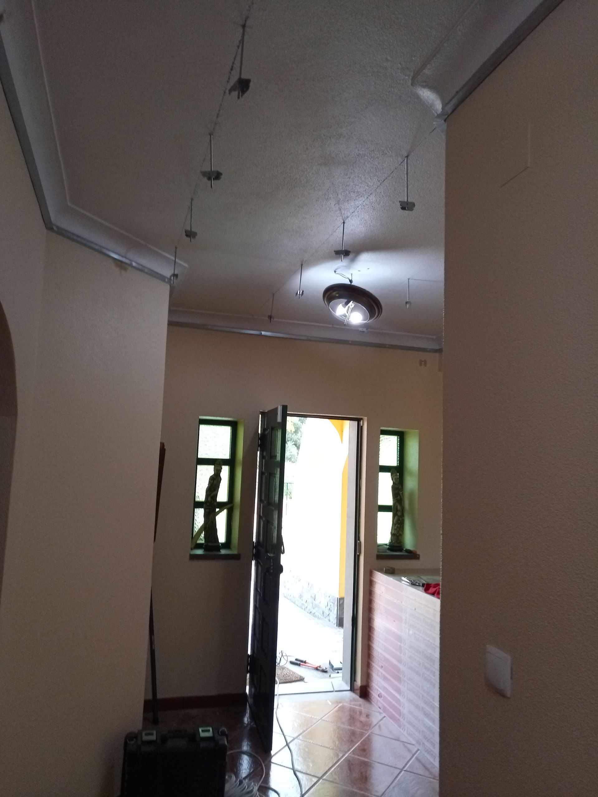 Corredor com luzes no teto, porta aberta para o exterior, janelas com moldura verde, paredes bege e piso de azulejo.