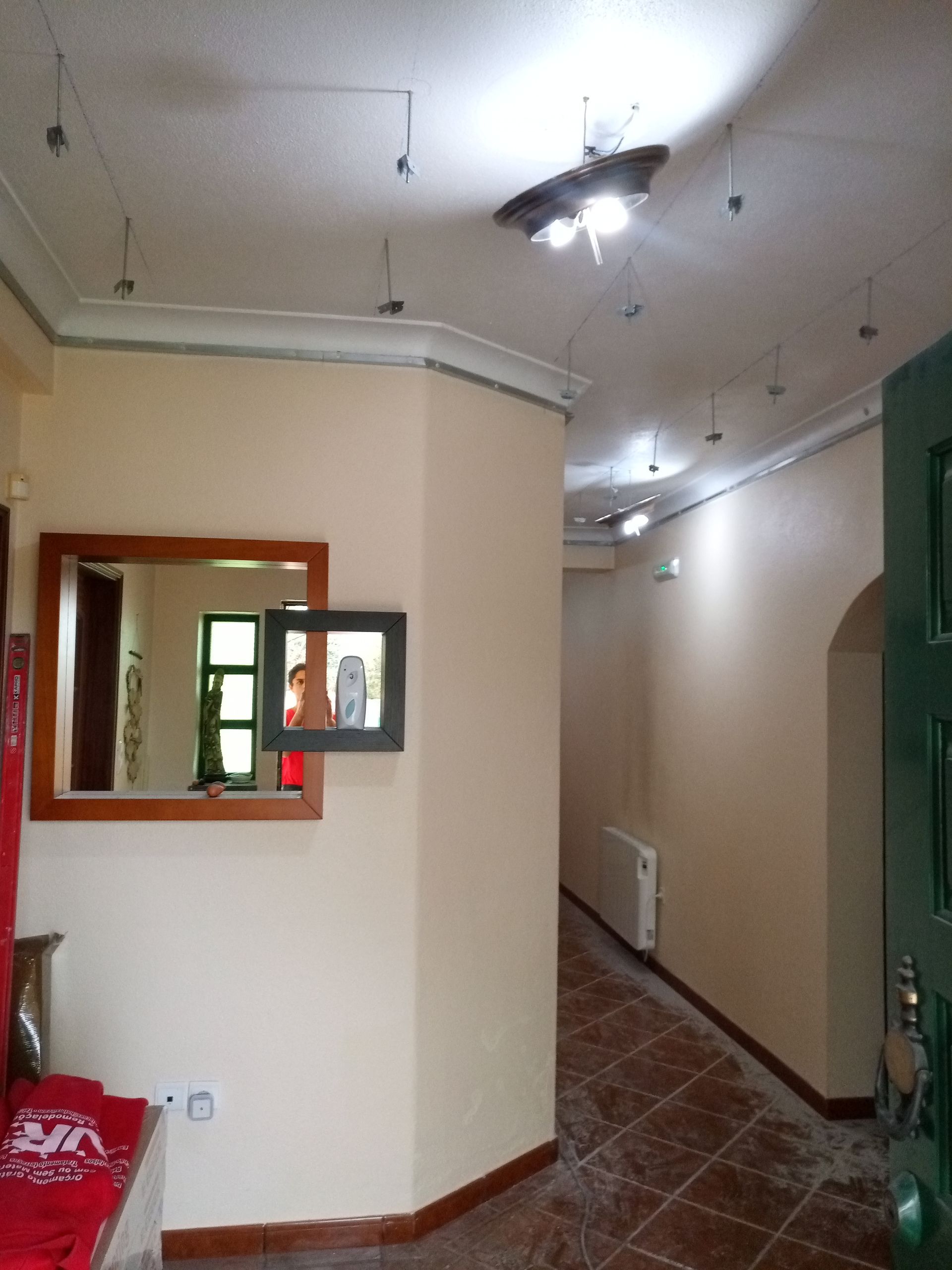 Corredor com paredes bege, piso de azulejo marrom e pequenas luzes. Um espelho reflete uma pessoa.