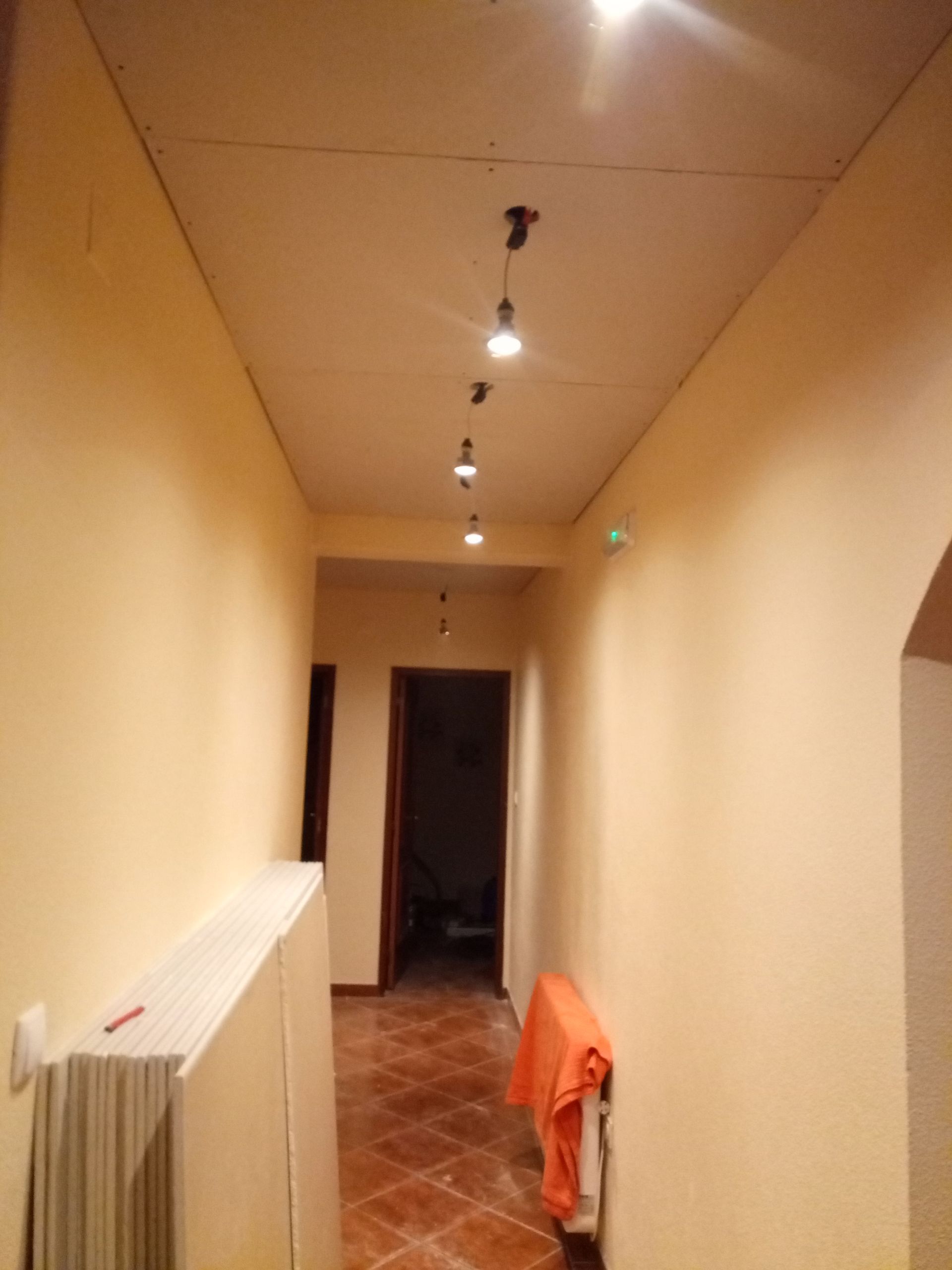 Corredor estreito com luzes no teto, paredes cor creme, piso de azulejo e portas abertas em intervalos regulares.