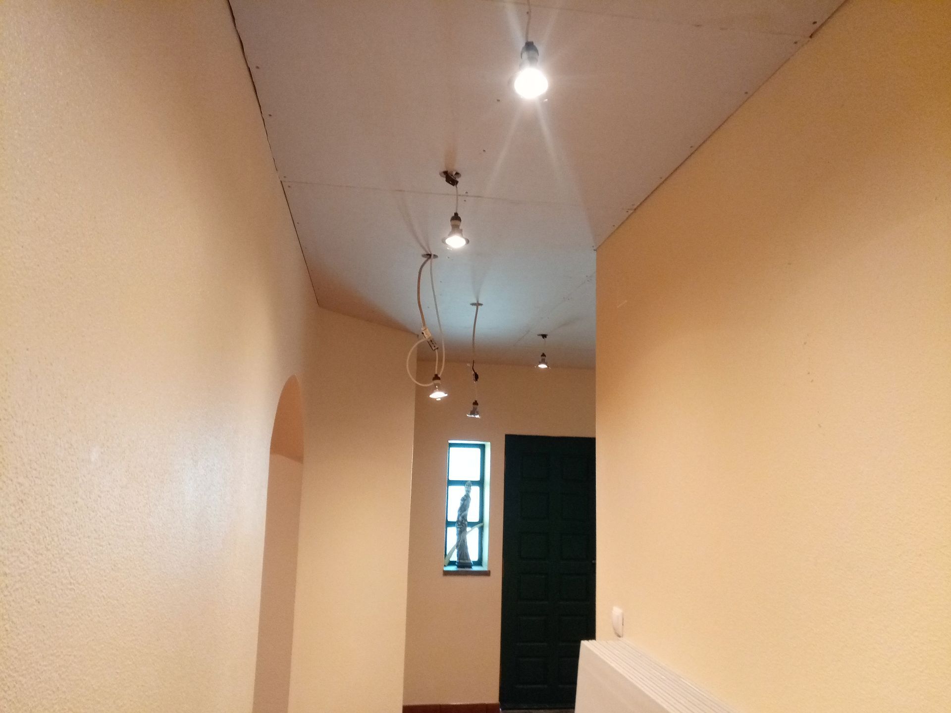 Longo corredor com paredes bege, teto branco e lâmpadas pendentes que leva a uma porta e janela verde-escuras.