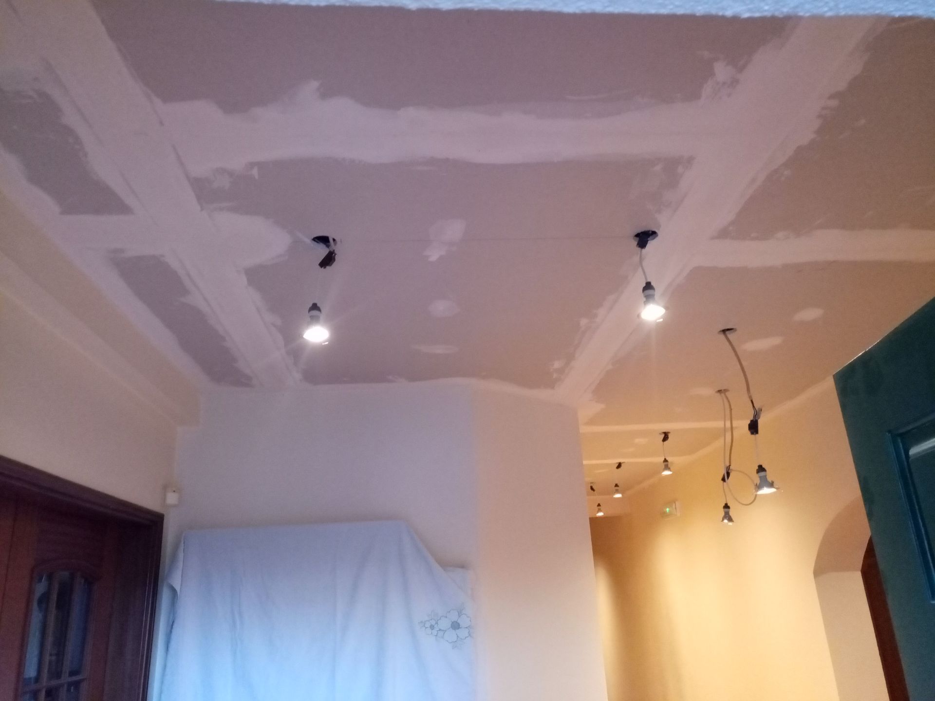 Teto com trabalho de drywall em andamento; áreas embutidas, iluminação em trilho, paredes cor creme e um lençol branco cobrindo algo.