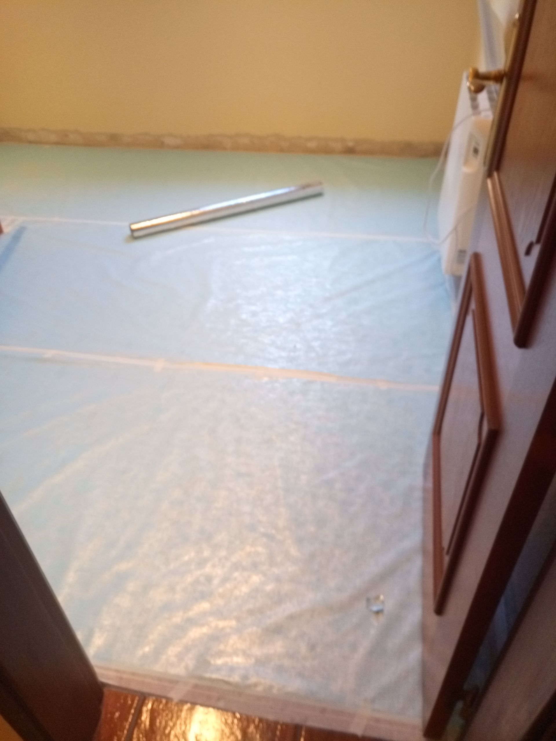 Sala com piso coberto por plástico azul, rolo prateado e porta aberta à direita.