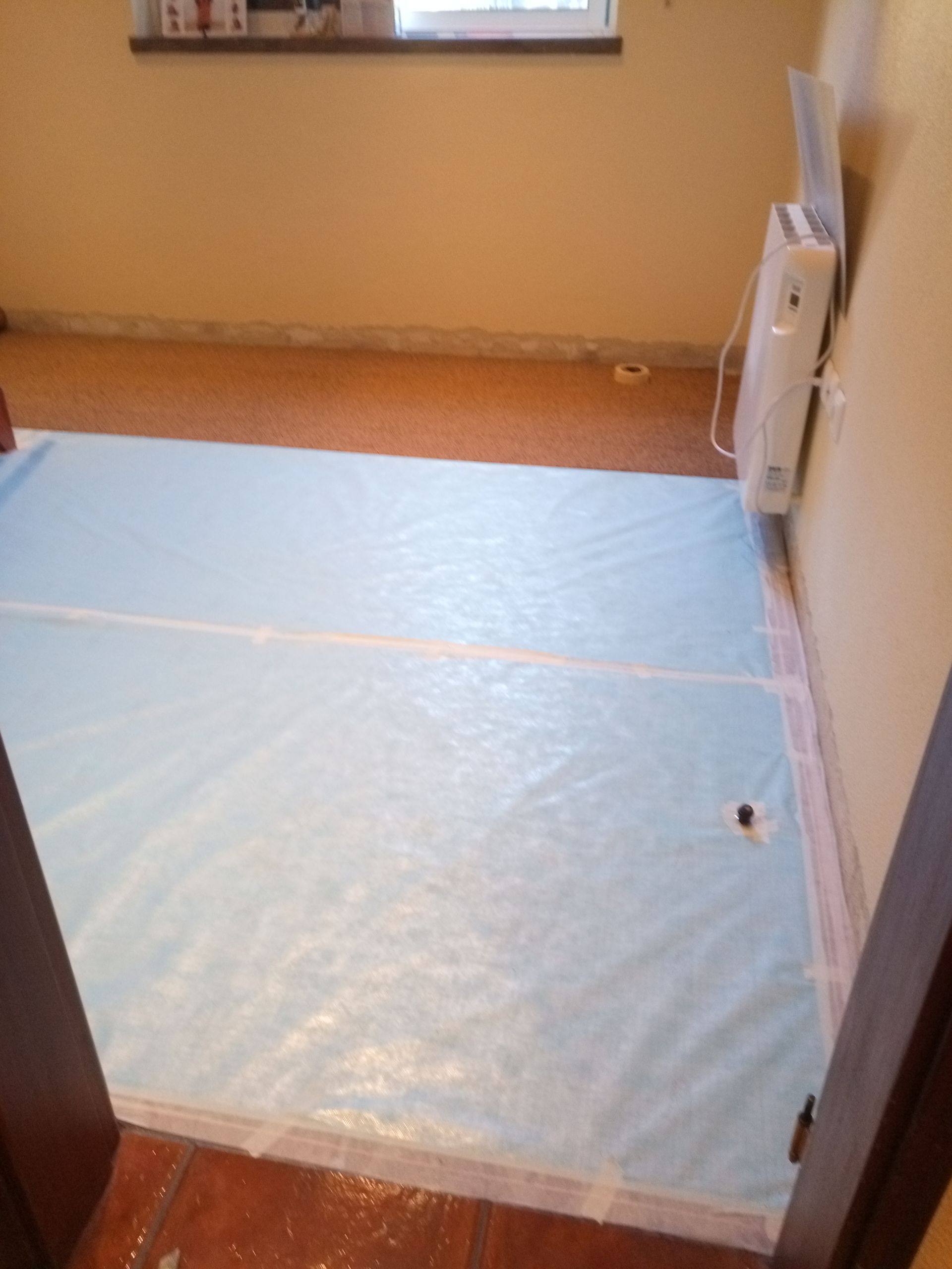 Lona plástica azul cobrindo o chão de um cômodo, perto de um aquecedor em uma parede bege, provavelmente para instalação de piso.