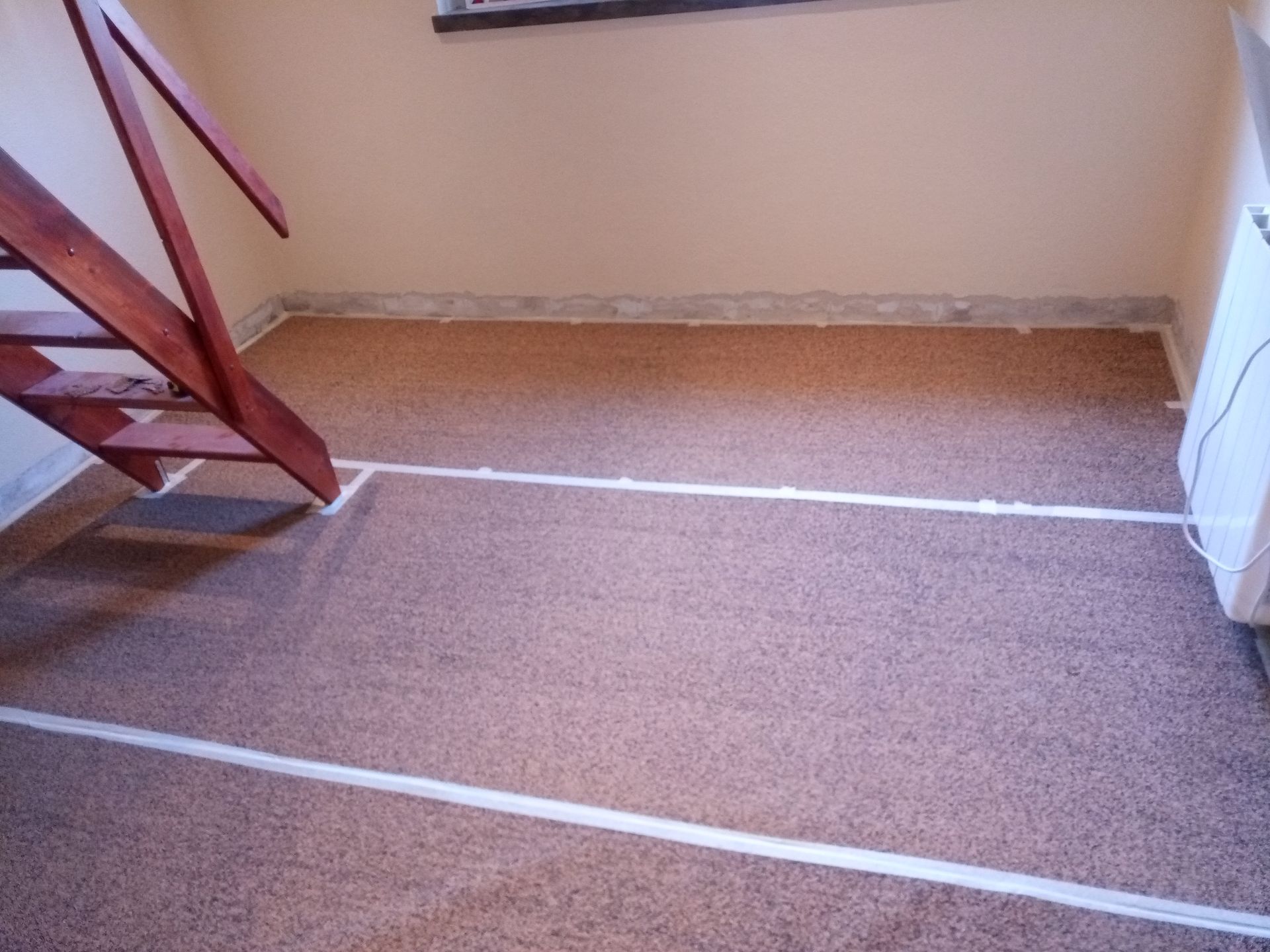 Piso de cortiça marrom com linhas brancas marcadas com fita adesiva, adjacente a uma escada de madeira e uma parede.