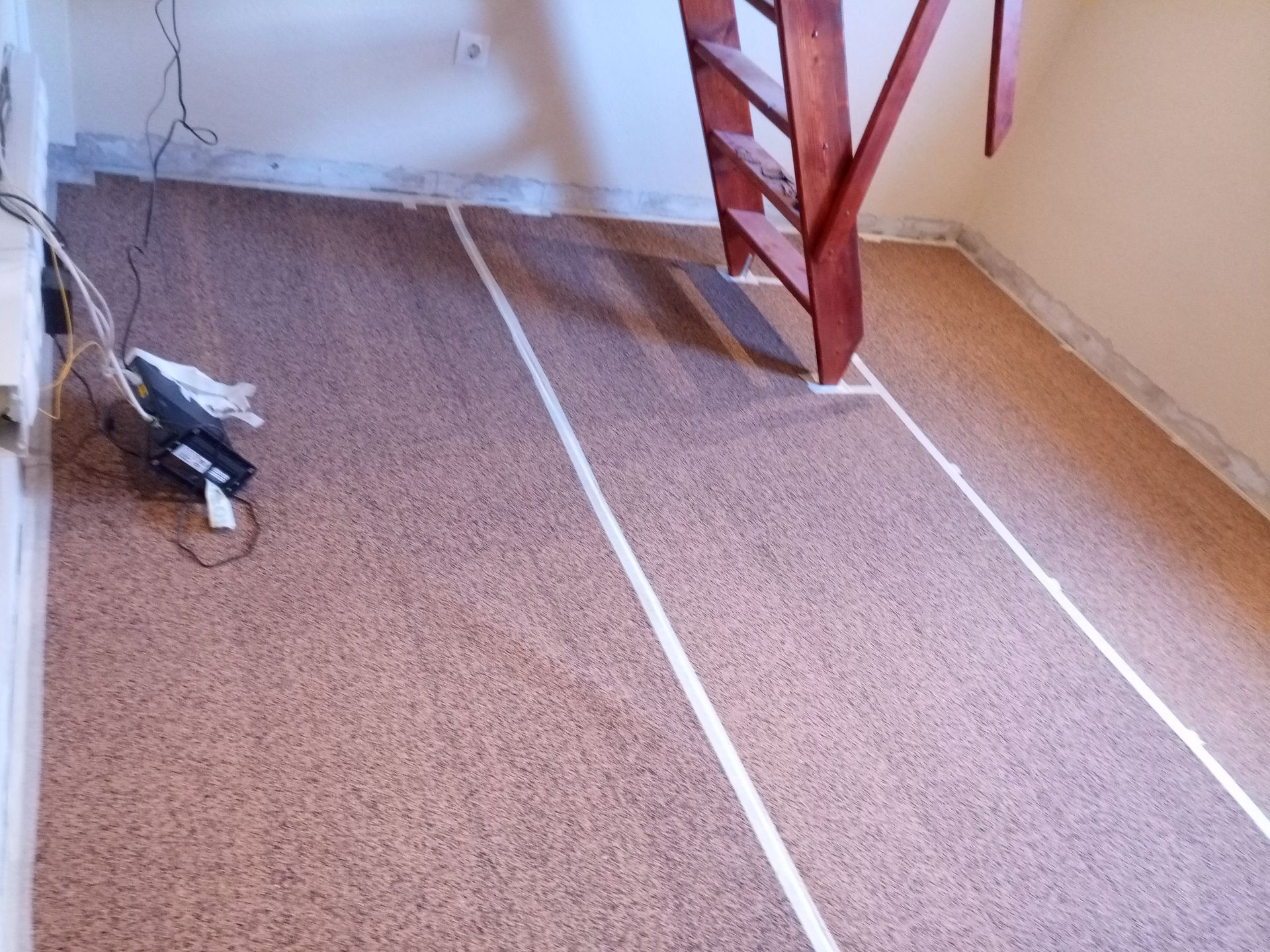 Piso de cortiça marrom com linhas de fita adesiva branca, próximo a uma escada de madeira e paredes.