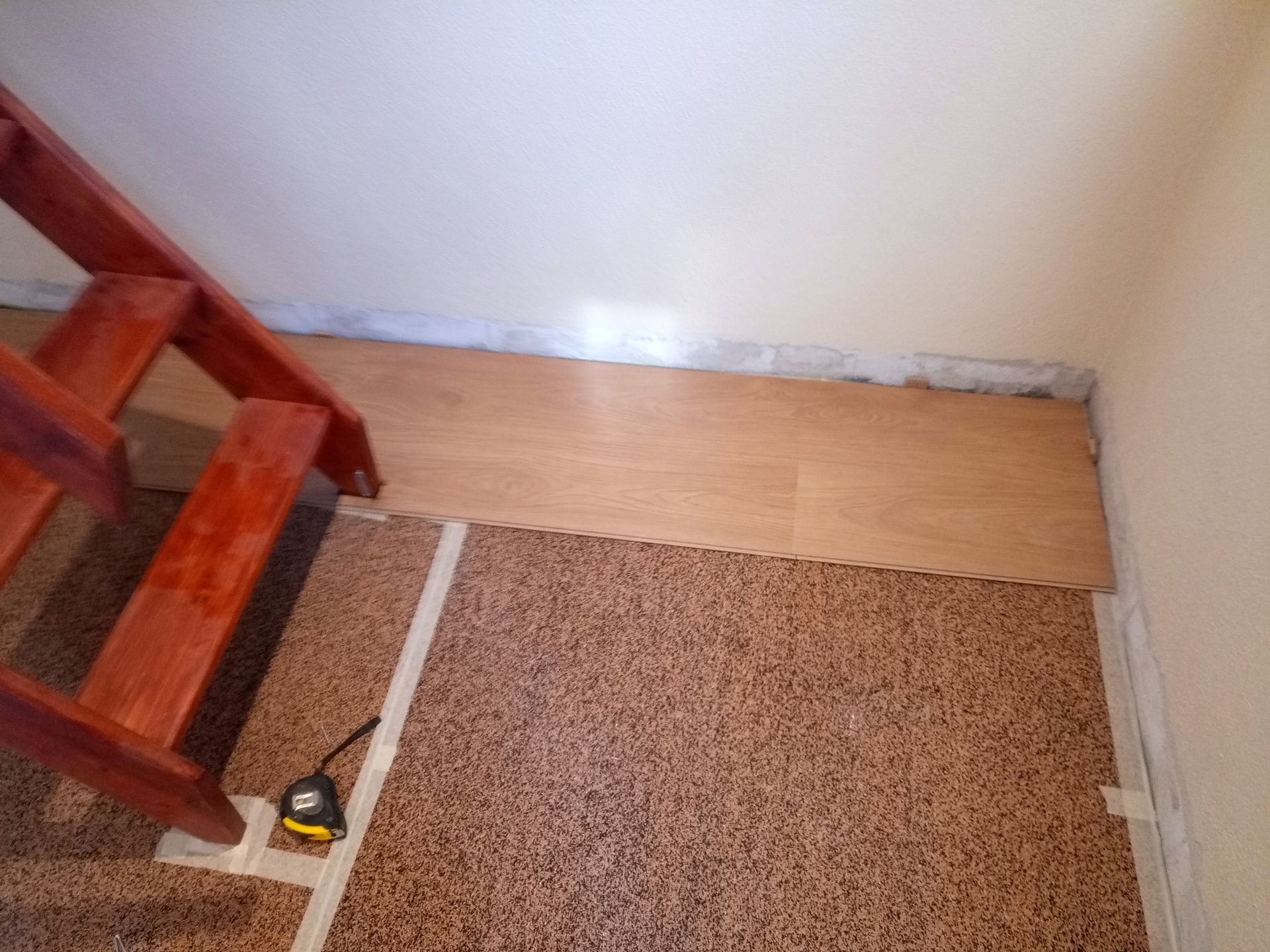 Piso de madeira instalado ao lado de uma escada e piso marrom com manchas, fita métrica e trena visíveis.