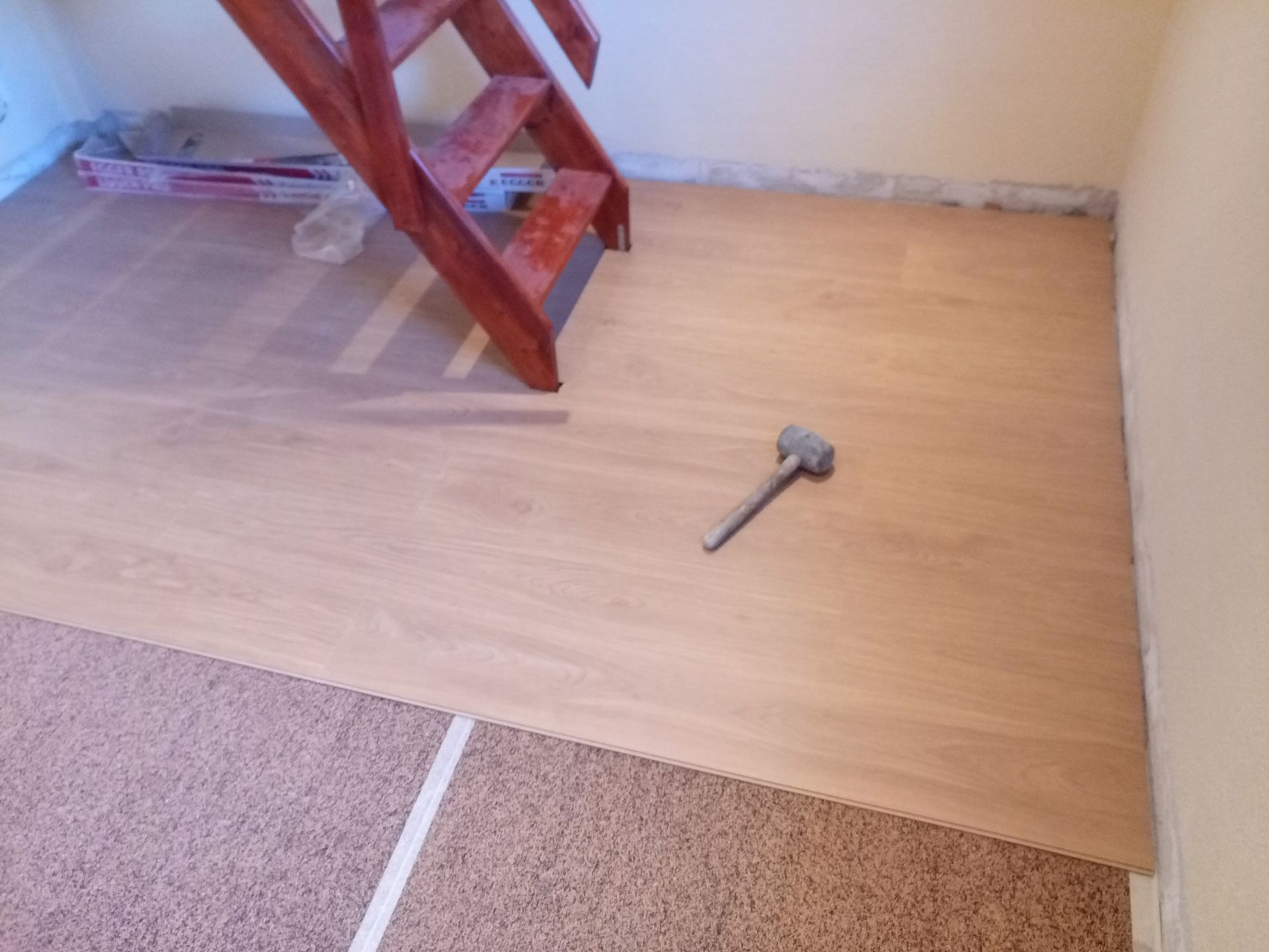 Piso de madeira sendo instalado próximo a uma escada vermelha. Um martelo está apoiado no chão.