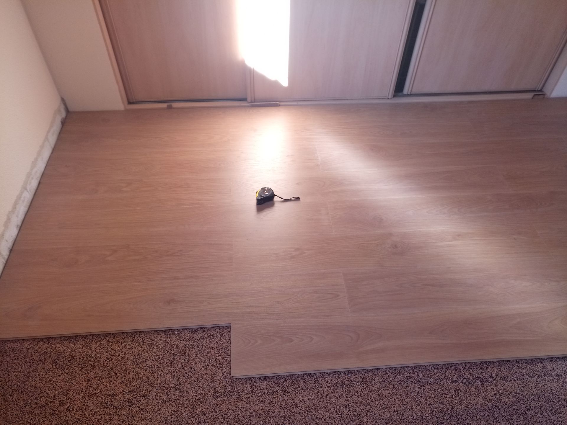 Piso laminado marrom sendo instalado, fita métrica sobre a superfície. Carpete visível por baixo.