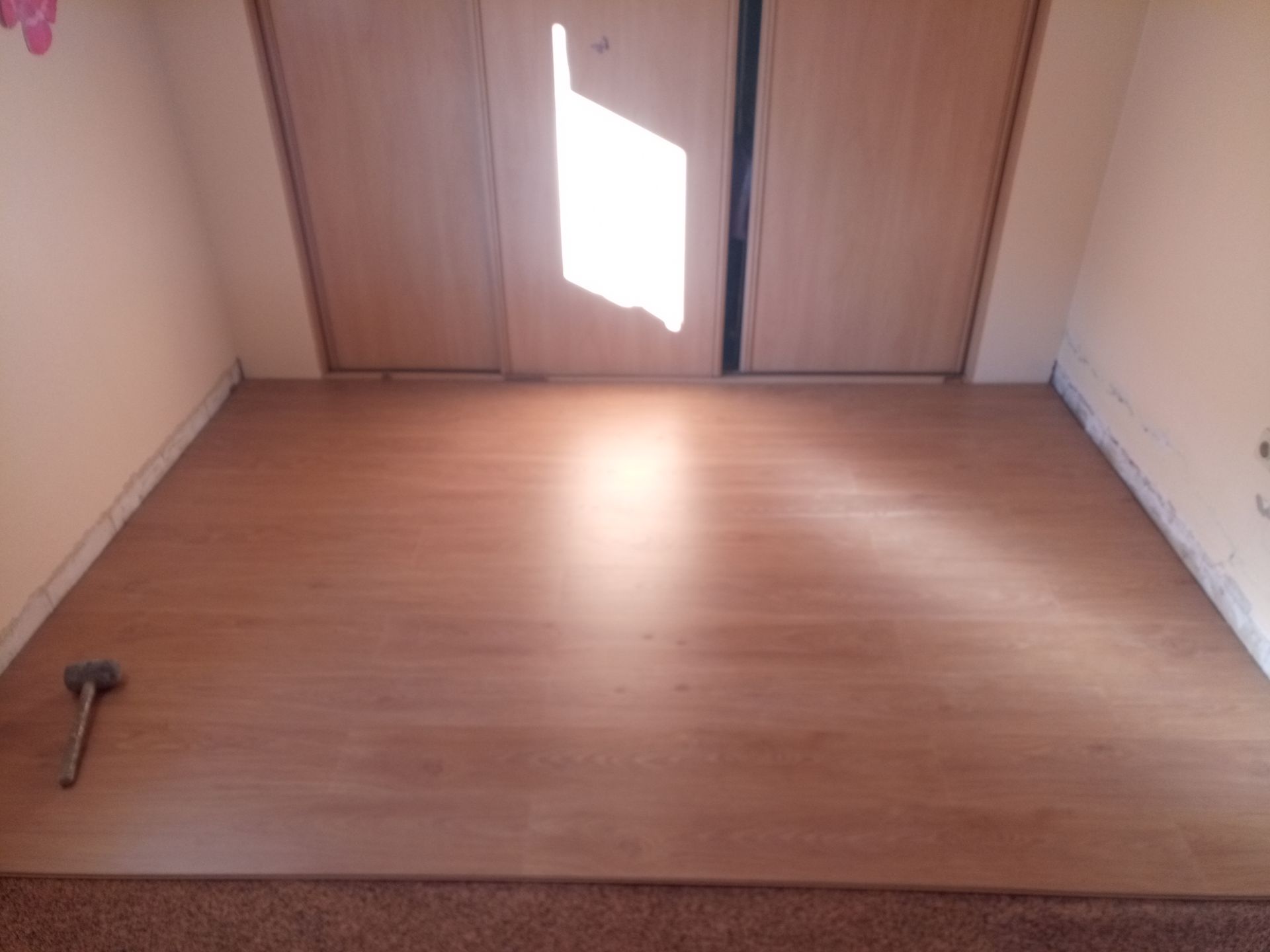 Piso de madeira em um quarto com duas portas de armário fechadas, e um martelo visível no chão.