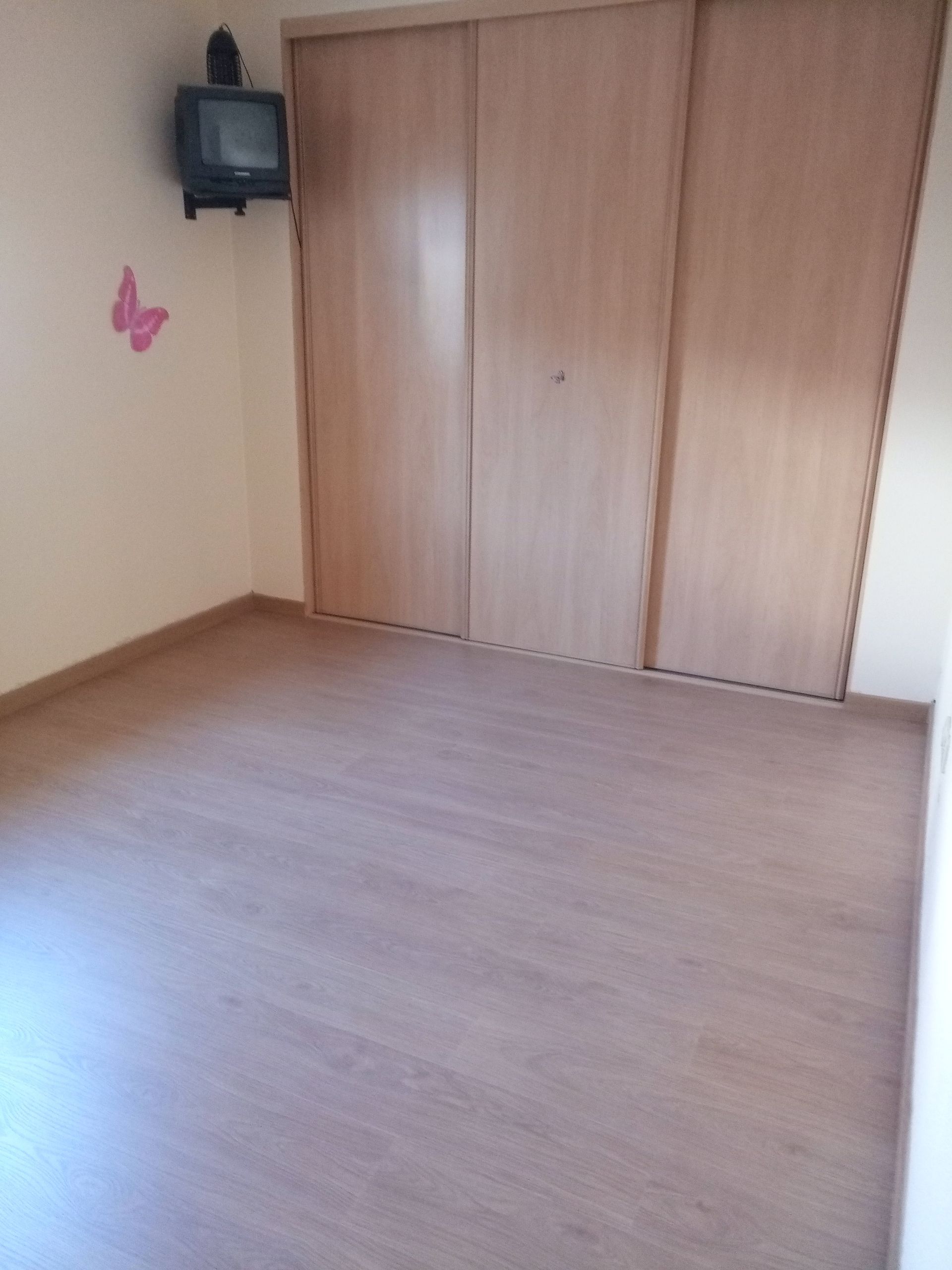 Quarto vazio com piso que imita madeira, paredes claras e um grande armário. Uma pequena TV está fixada na parede.
