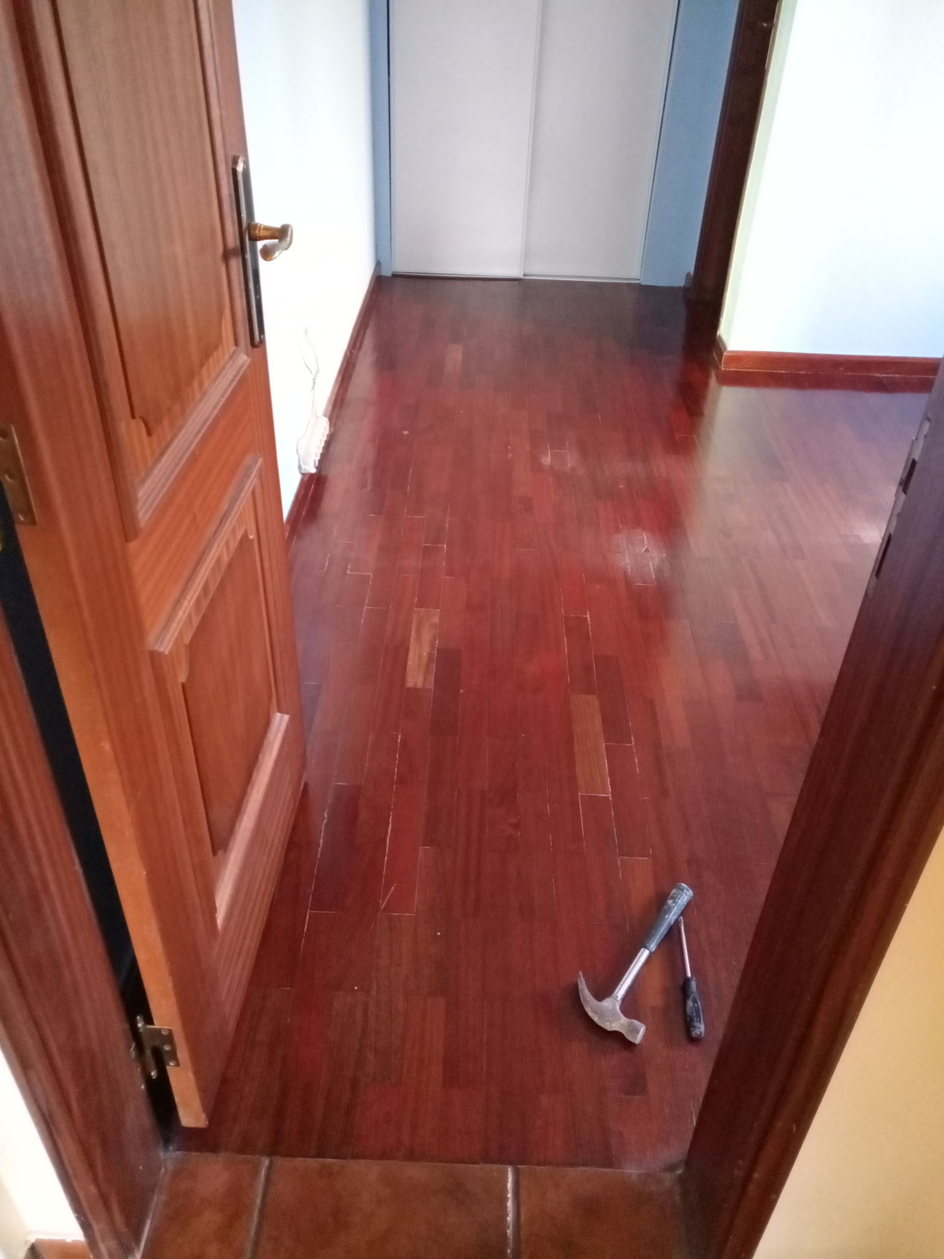 Piso de madeira em um corredor com uma porta marrom aberta. Um martelo está no chão.