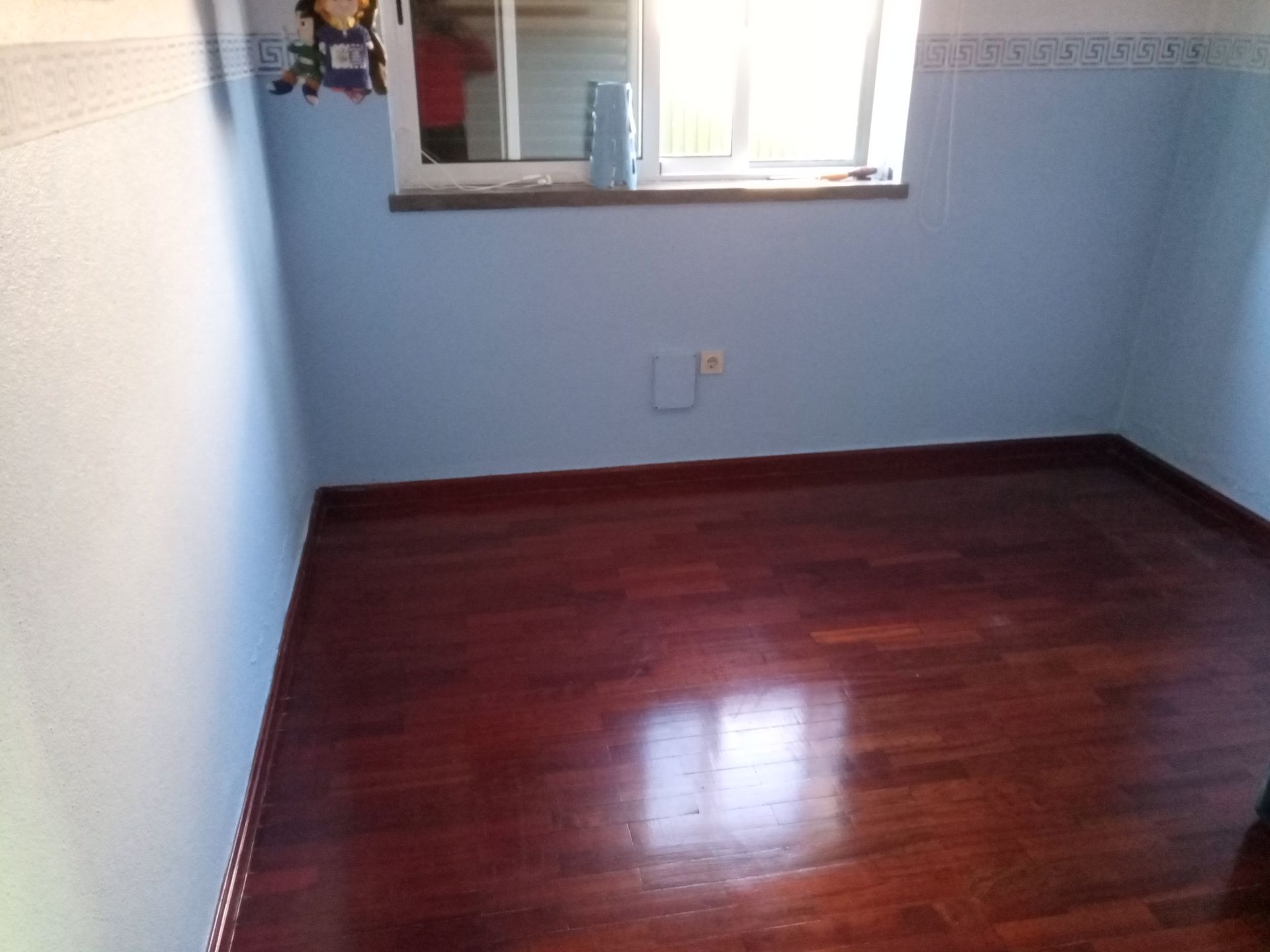 Quarto vazio com paredes azuis, piso de madeira escura e uma janela.