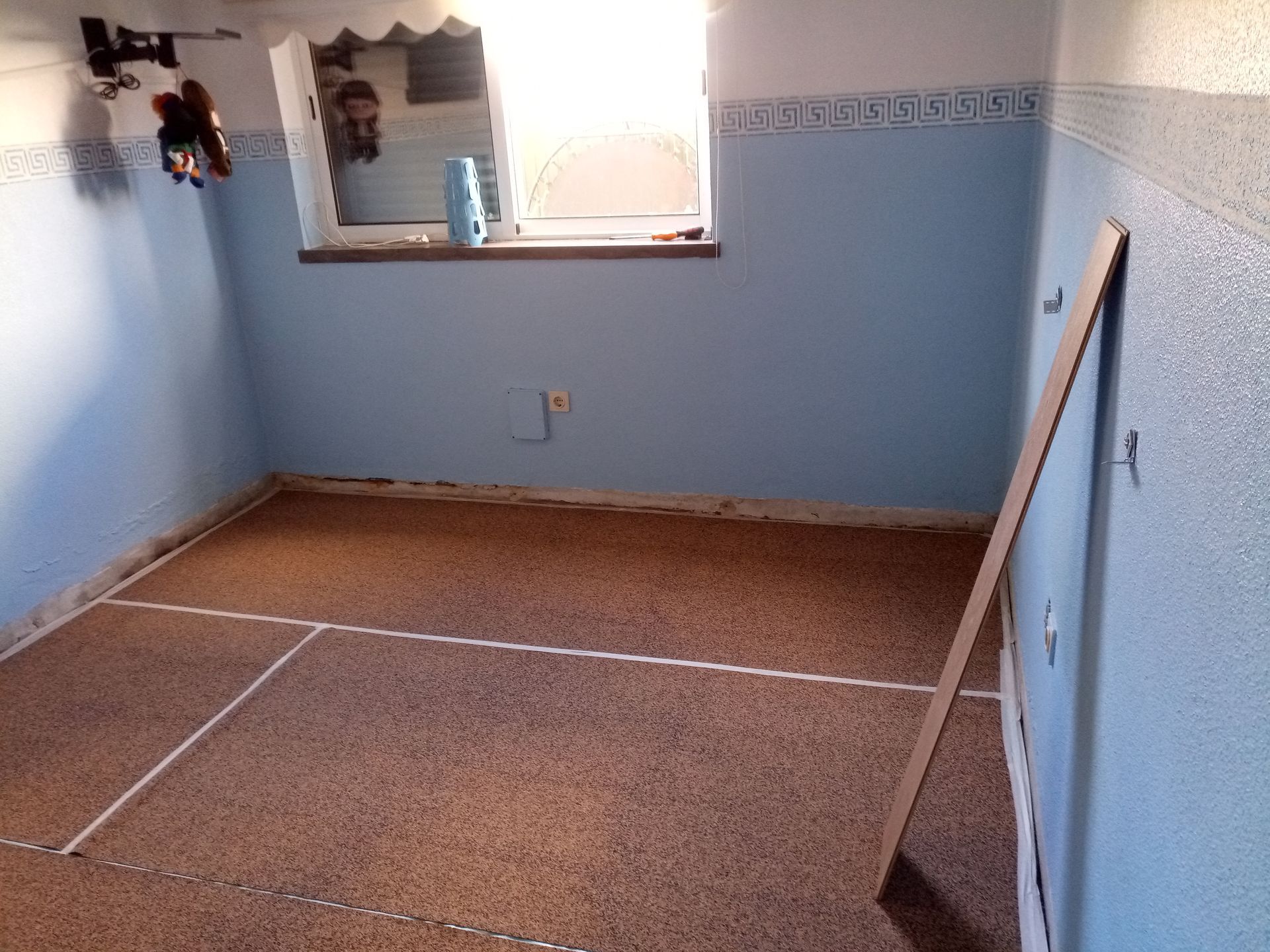 Um quarto com paredes azuis, uma janela e piso de cortiça com linhas brancas demarcando as seções.