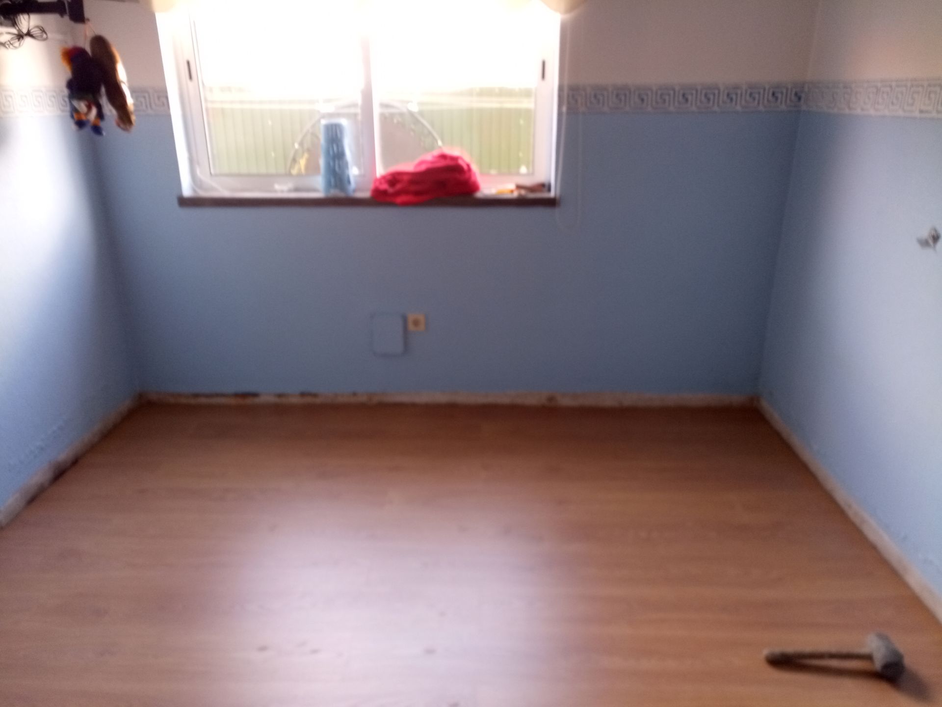 Quarto vazio com paredes azuis, uma janela e piso de madeira. Um martelo está sobre o chão.
