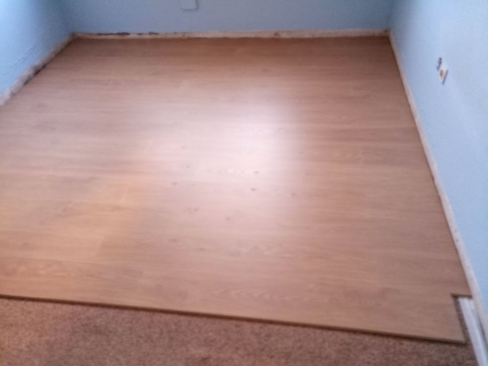 Piso laminado de madeira clara instalado em um cômodo com paredes azuis e faixa de carpete na borda.