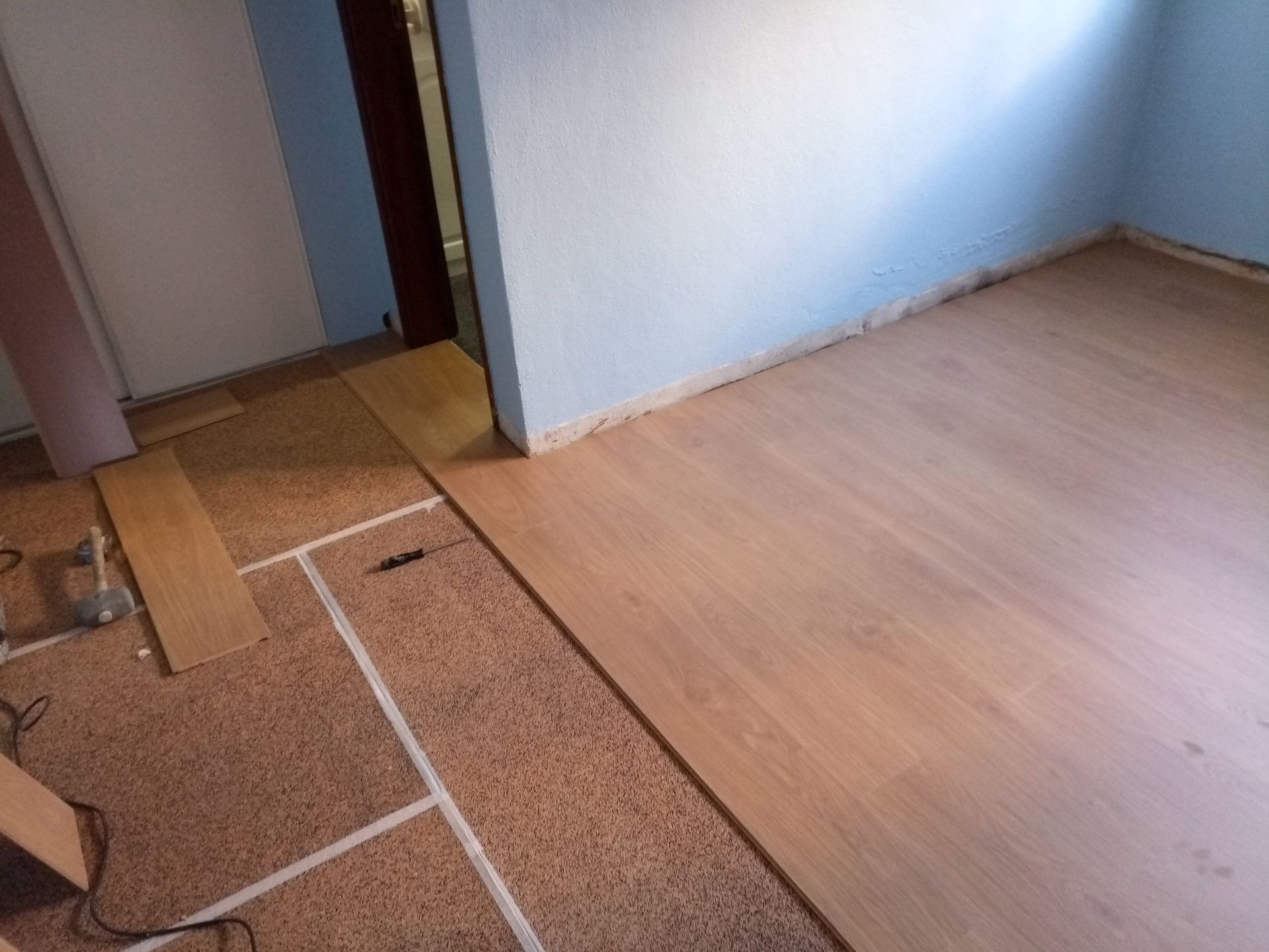 Instalação de piso de madeira maciça em um cômodo. As tábuas são marrom-claras; as paredes são azuis e brancas.