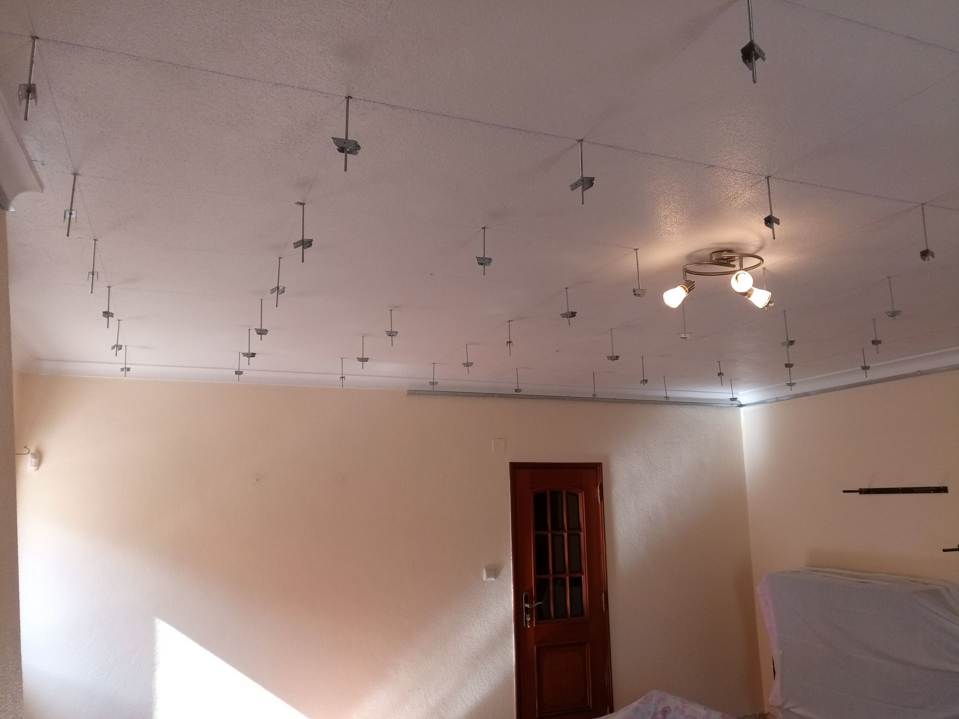 Teto branco com muitas barras de sustentação metálicas. O quarto possui uma porta e paredes cor creme.