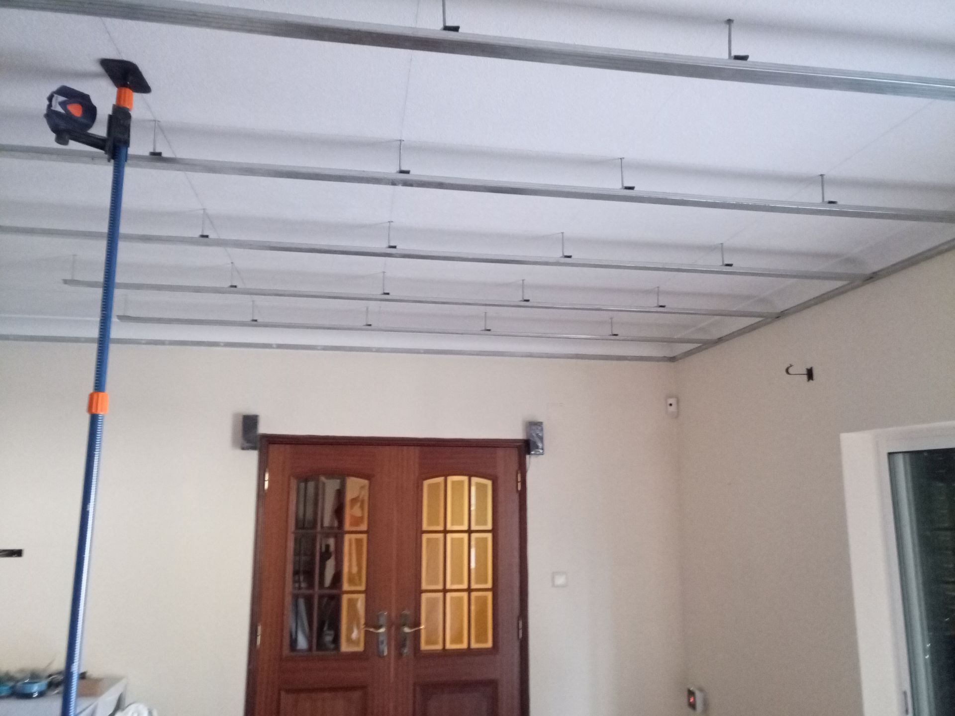 Estrutura de teto com armação metálica e fios de sustentação, em um cômodo com porta dupla e instrumento de medição.