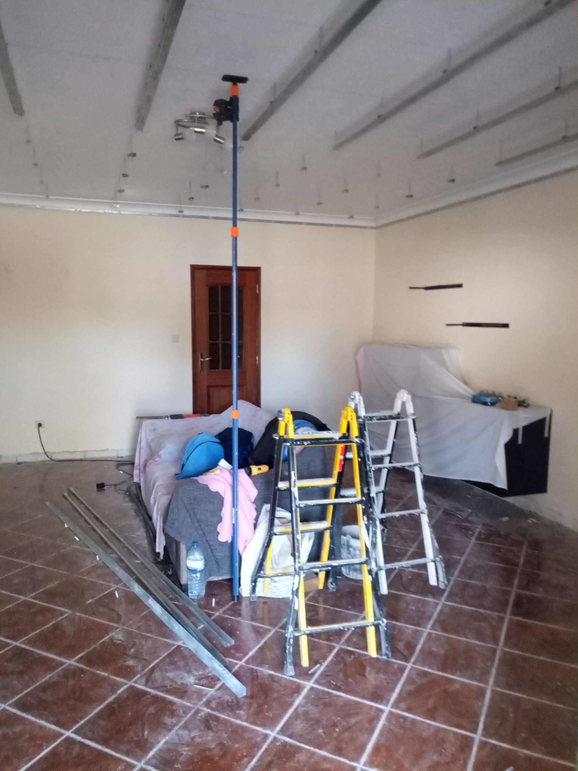 Quarto em reforma; poste de sustentação no teto, escadas, sofá coberto com tecido, ferramentas, piso de azulejo marrom.