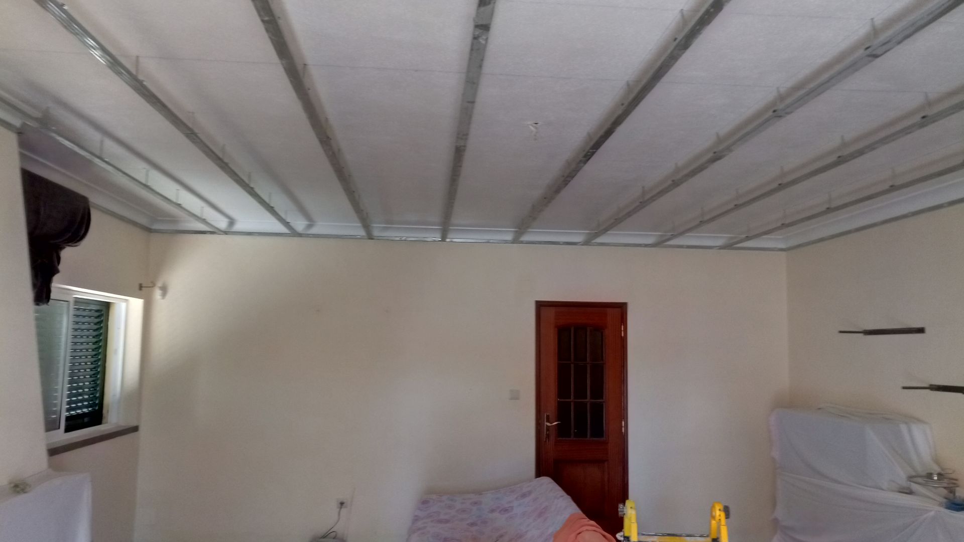 Vista interior de um cômodo, com teto com vigas metálicas e painéis brancos, porta, janela e paredes pintadas.