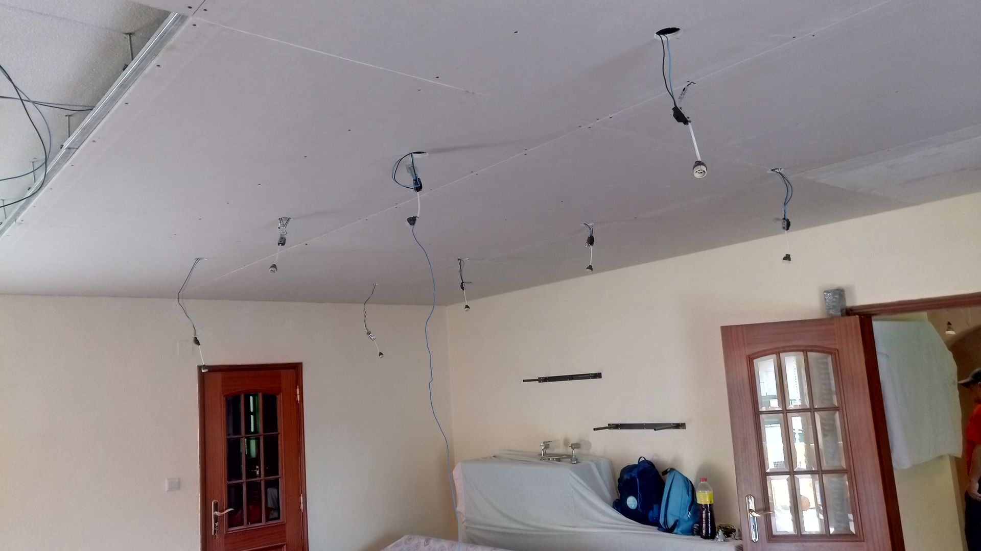 Teto com fiação exposta e luminárias pendentes em um cômodo com porta fechada, sofá e paredes cor creme.