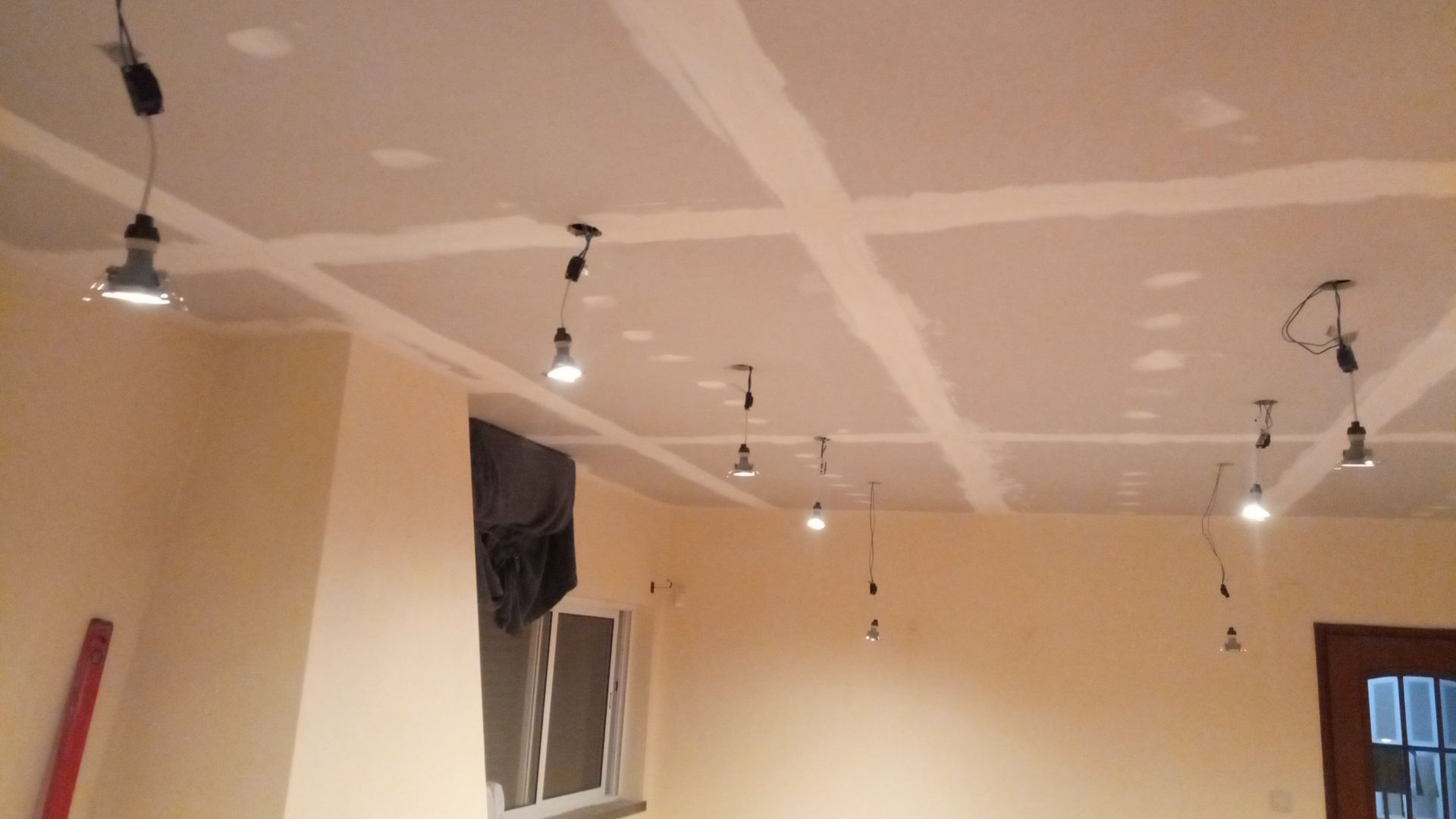 Vista interna de um cômodo com teto de gesso parcialmente acabado e luminárias aparentes penduradas. As paredes são pintadas de bege.