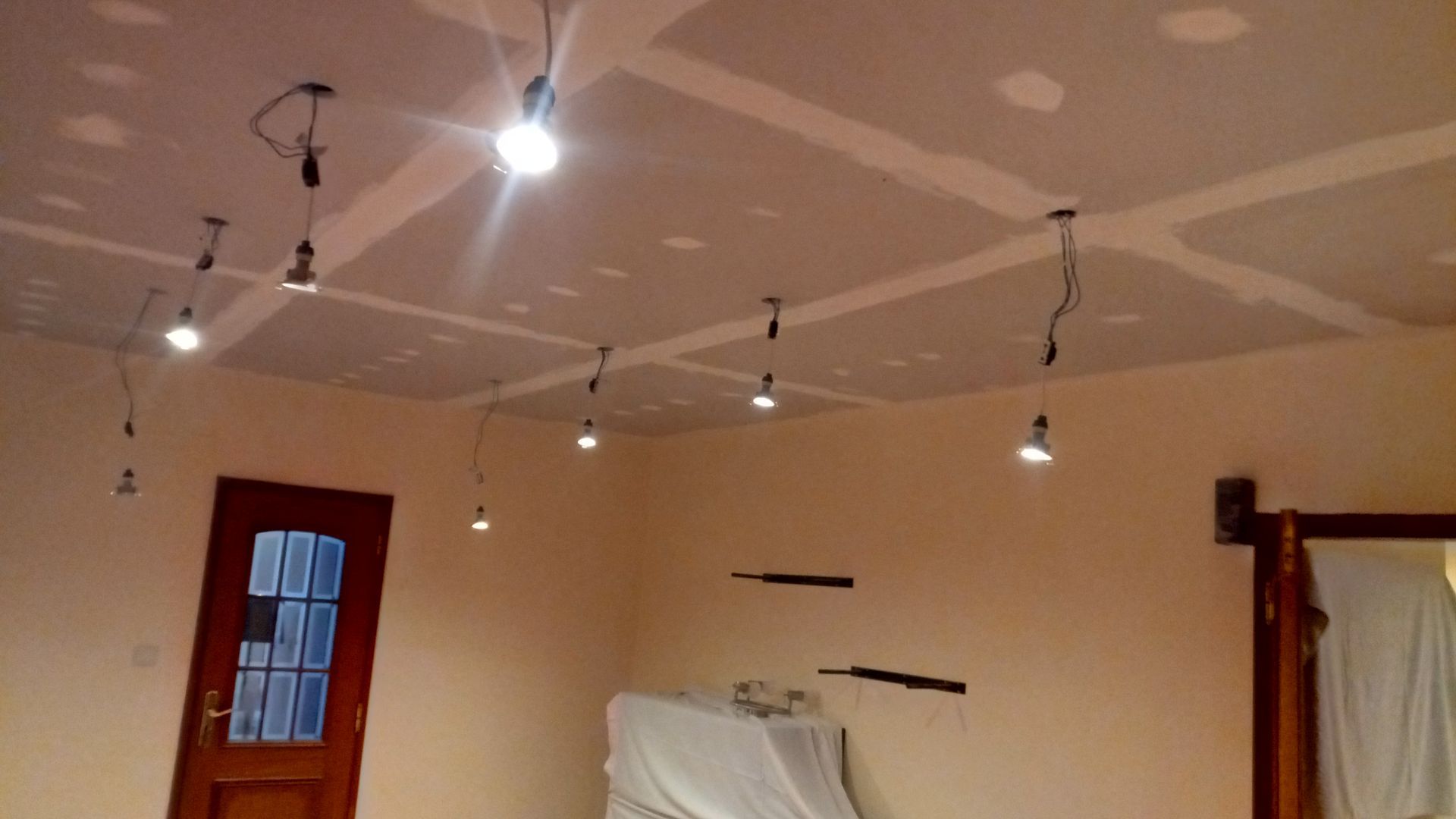 Vista interior de um cômodo com lâmpadas expostas penduradas no teto. As paredes são bege e o teto está inacabado.