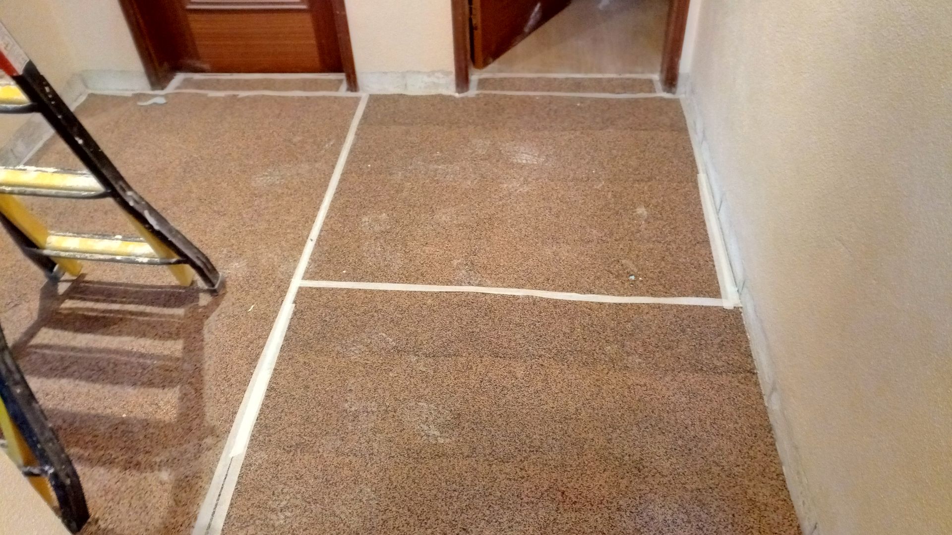 Piso marrom com manchas e linhas brancas demarcadas com fita adesiva, portas e uma escada de mão.