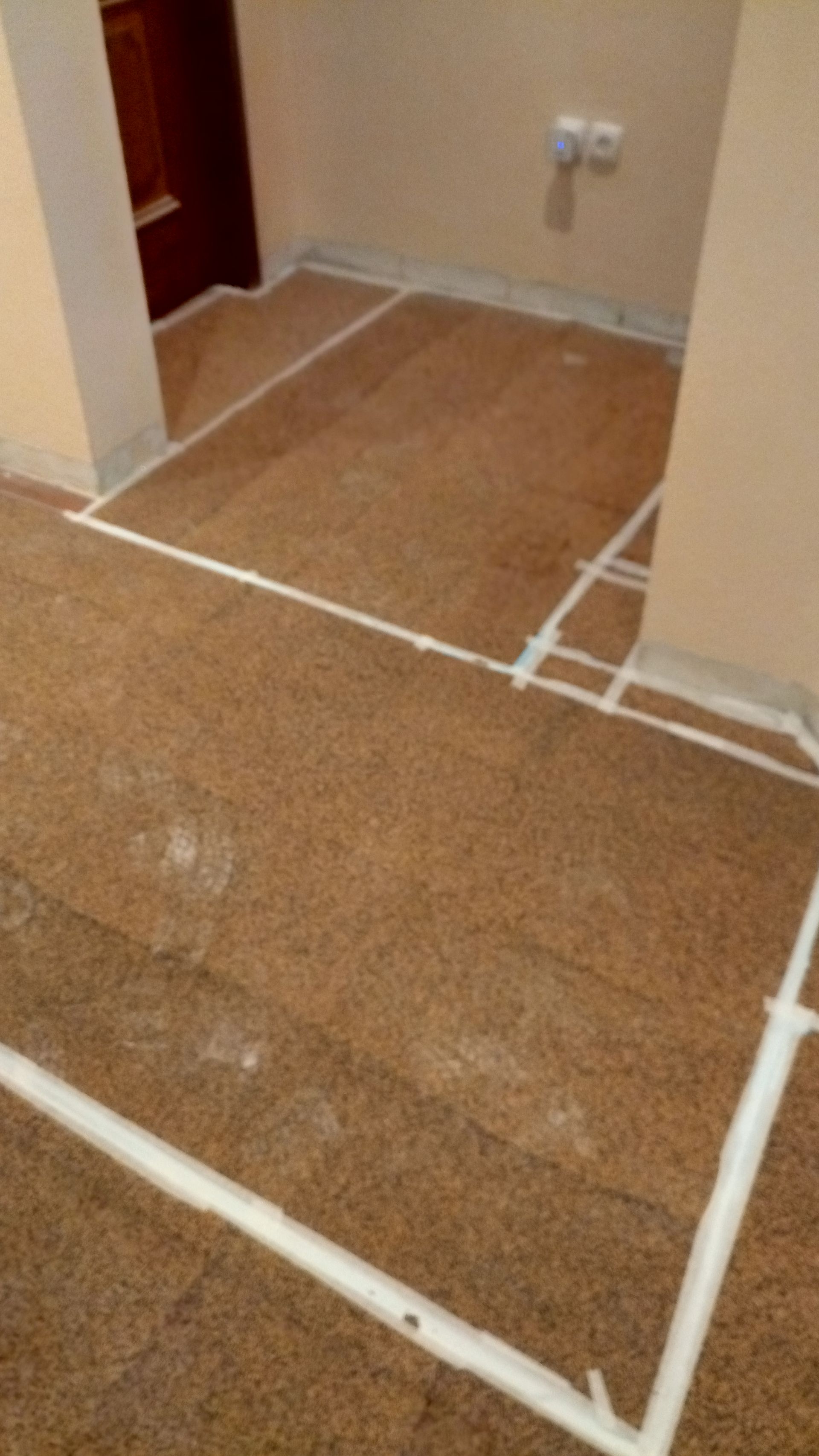 Piso marrom com fita adesiva branca demarcando as seções. Paredes bege e uma porta escura são visíveis.