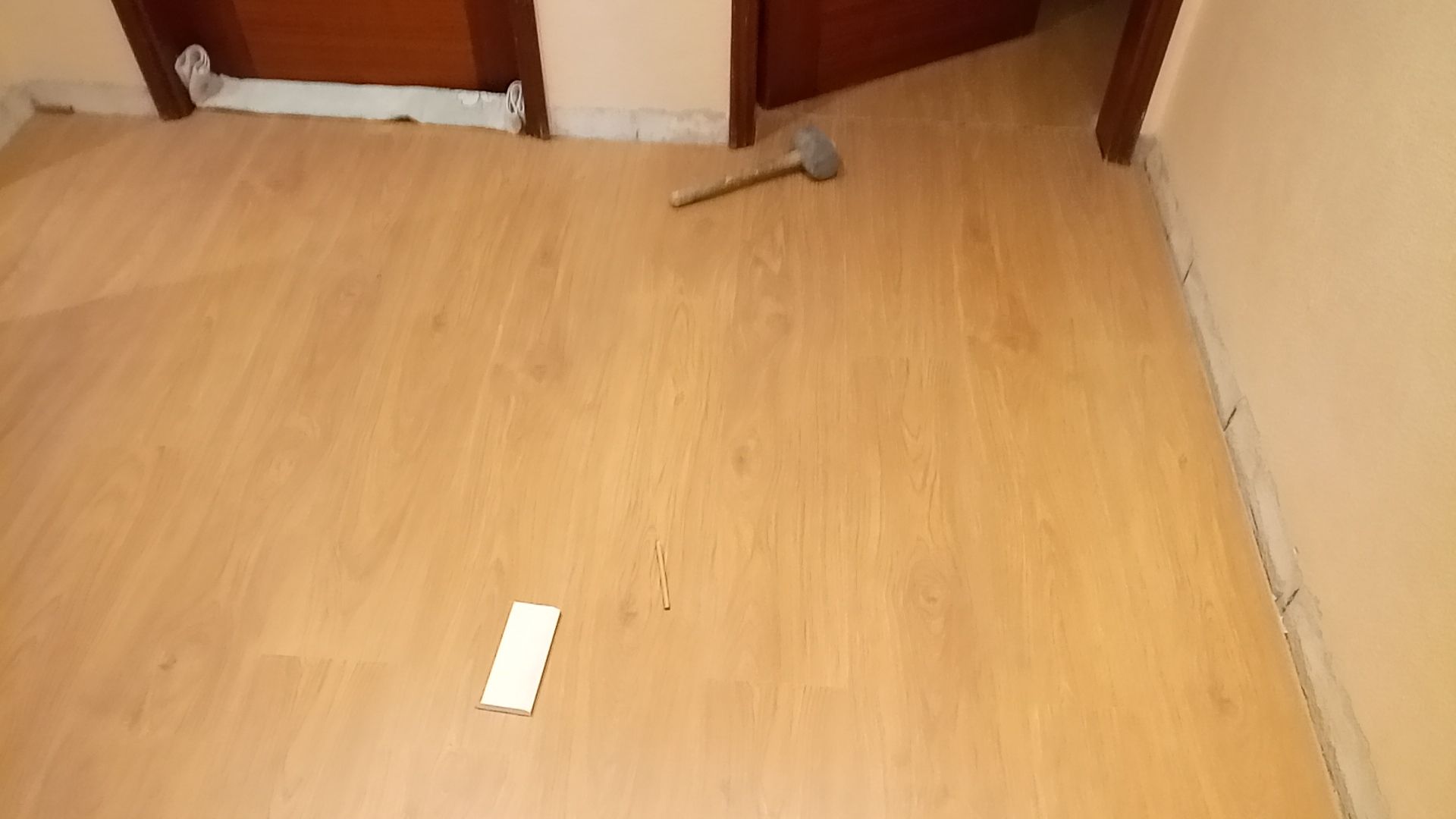 Piso de madeira clara com um martelo e um pedaço de piso em um cômodo vazio, ao lado de uma porta.