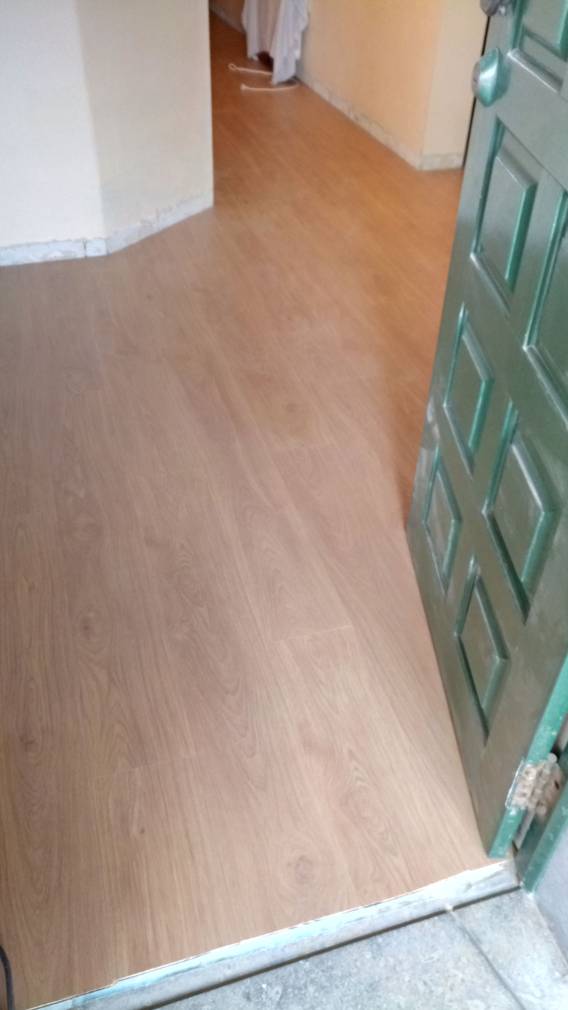 Piso de madeira que dá acesso a um cômodo. Porta verde à direita. Parede branca à esquerda.