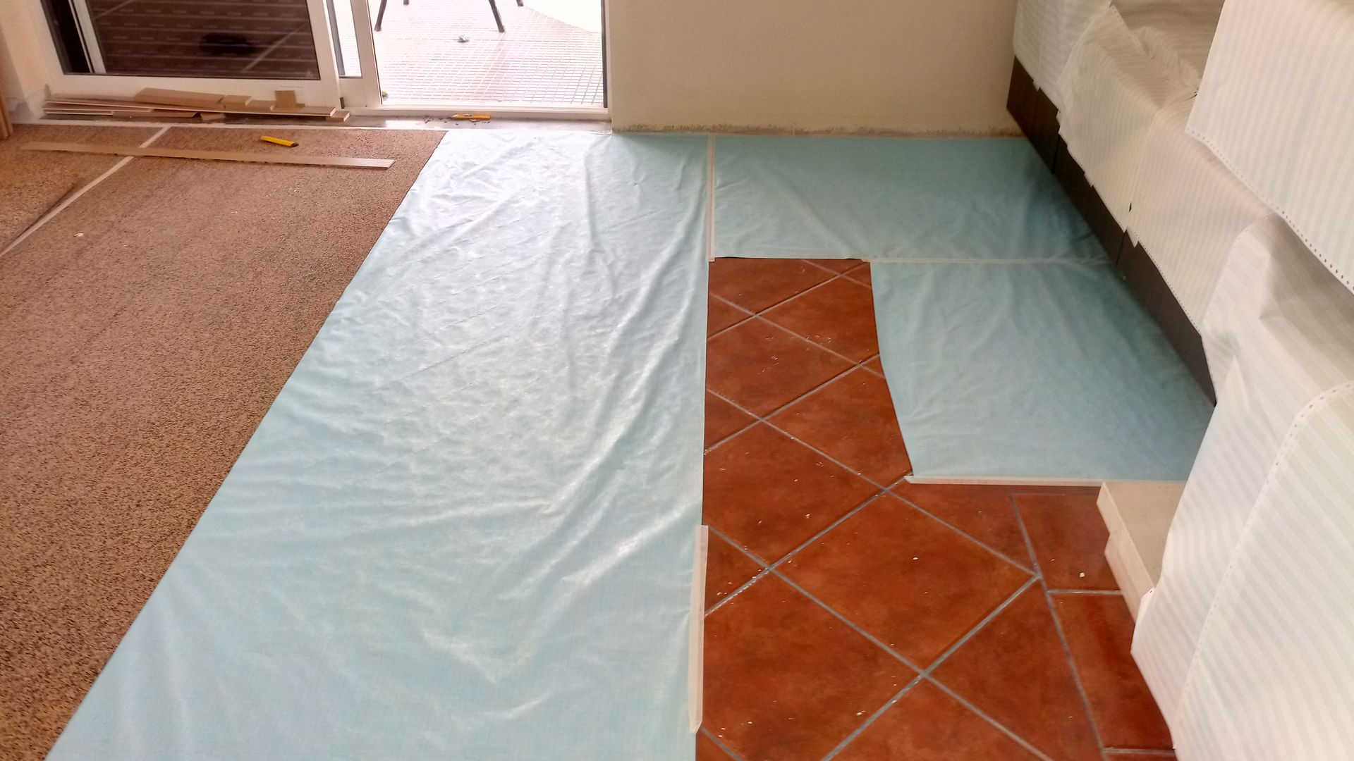 Chão coberto com lona plástica azul, ladrilhos de terracota e uma porta ao fundo.