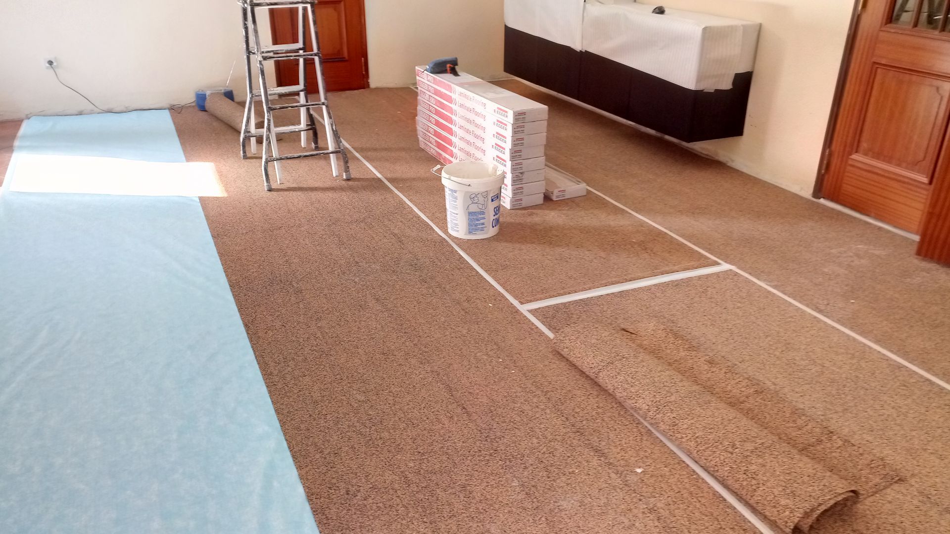 Instalação de piso de cortiça em andamento: tábuas marrons assentadas, subpavimento azul, escada e materiais sobre o piso claro.