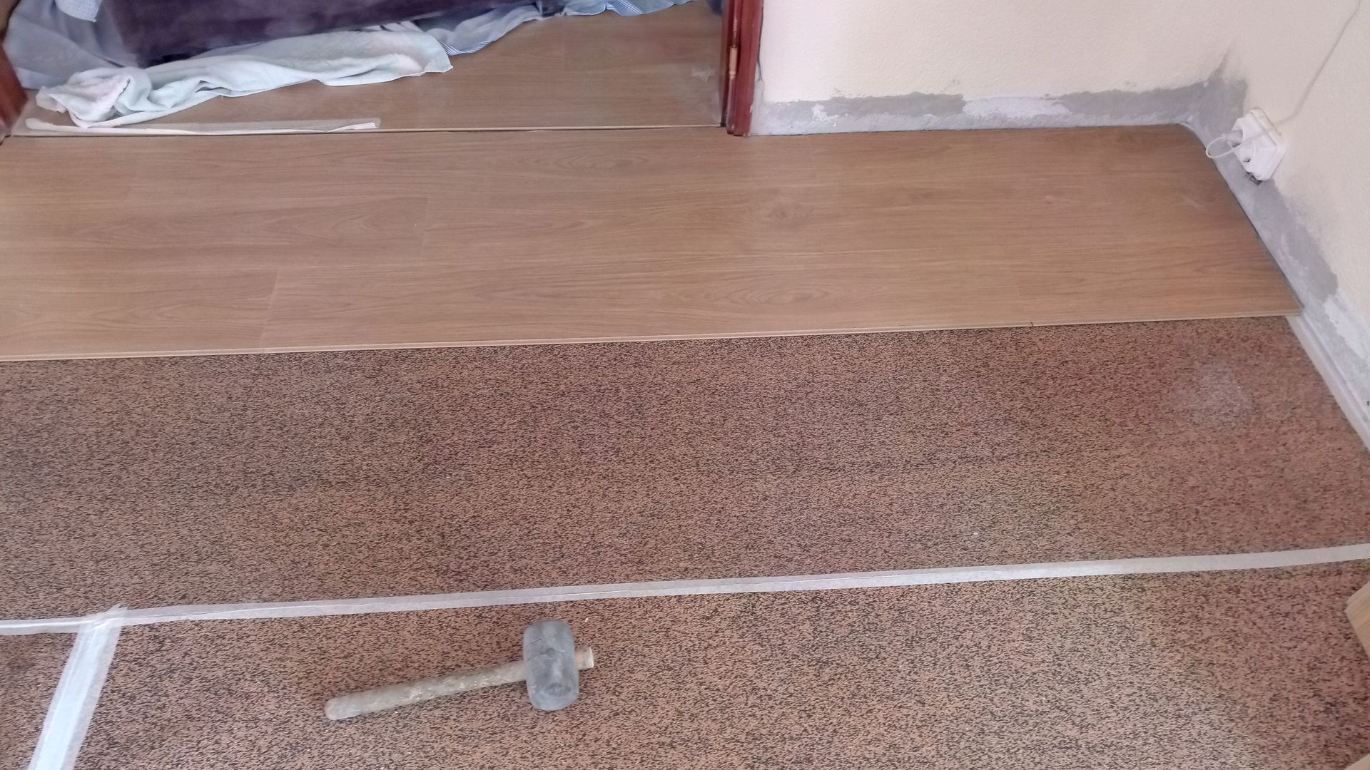 Instalação de piso em andamento: carpete marrom com tábuas de madeira sendo colocadas perto da porta. Martelo presente.