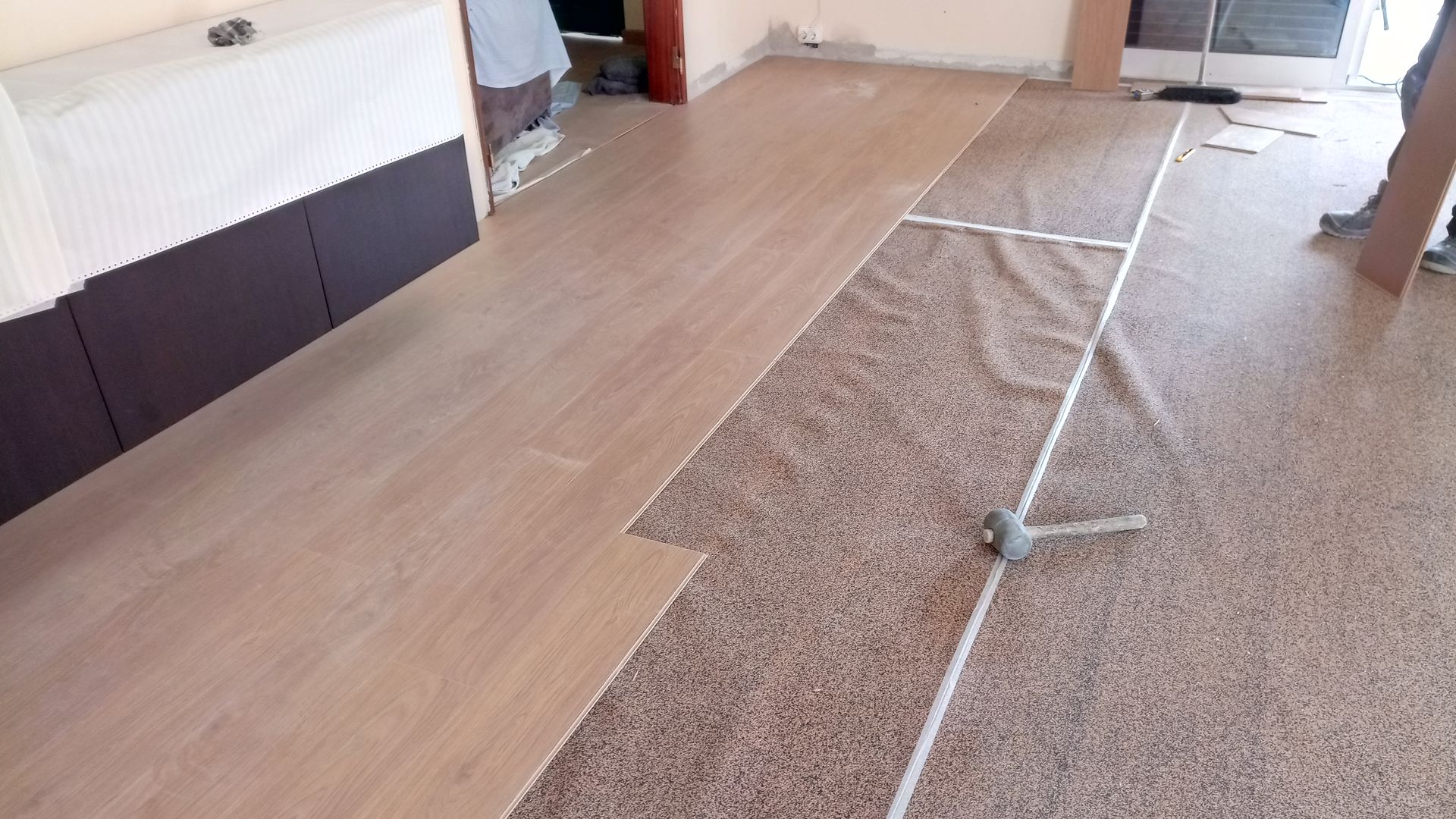 Instalação de piso laminado marrom claro sobre carpete existente com manchas em um cômodo.