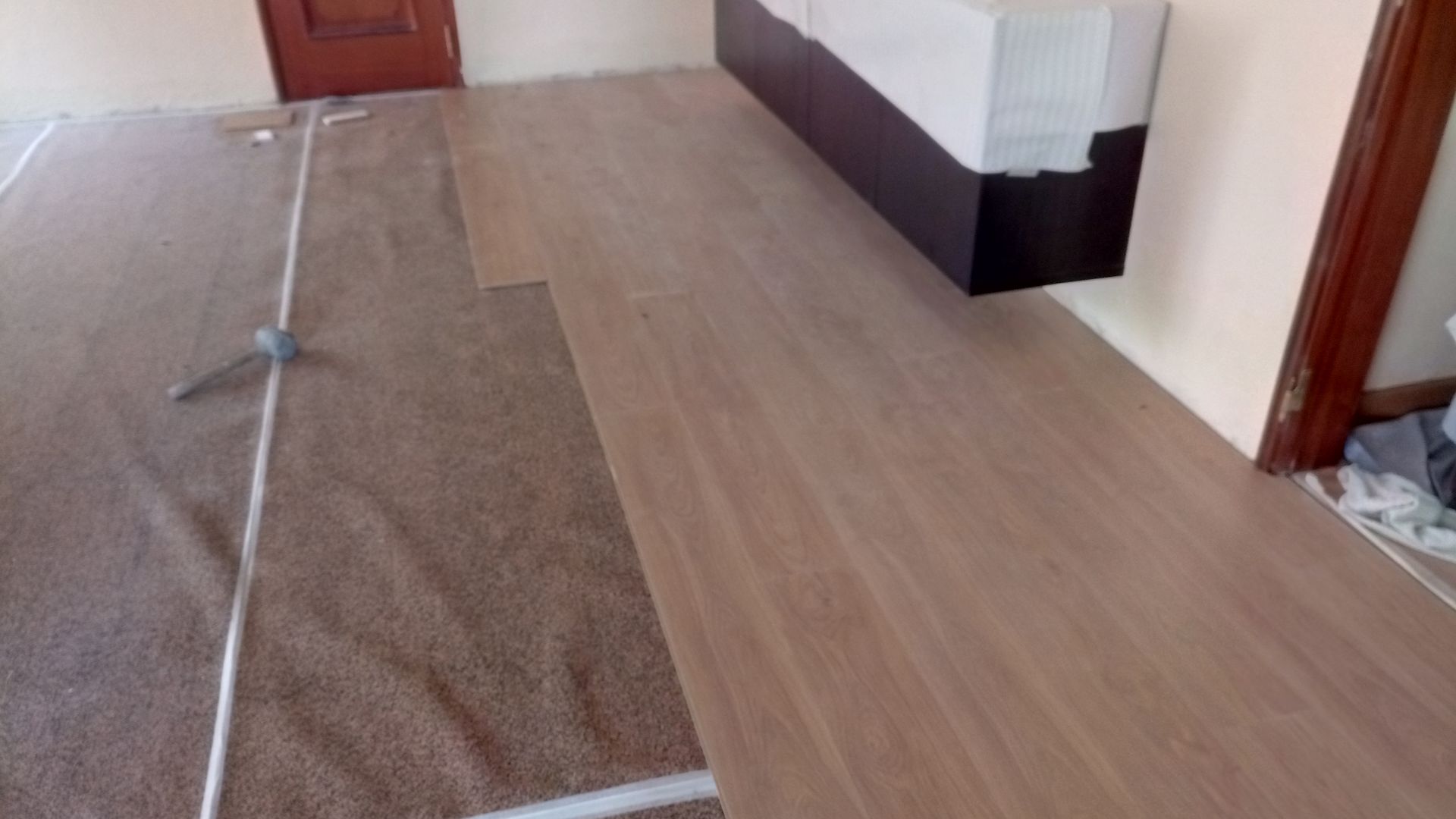 Quarto com carpete marrom sendo substituído por piso de madeira, ao lado de uma cama.