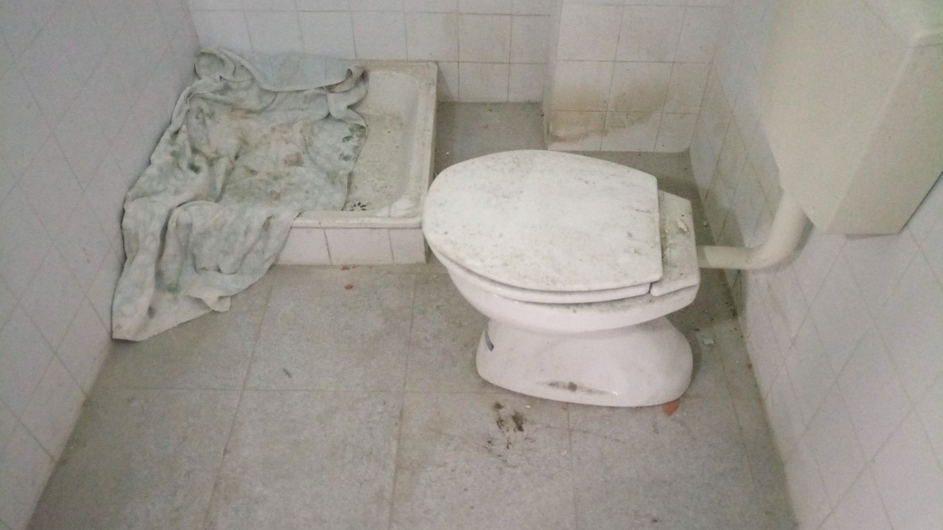 Vaso sanitário sujo e manchado em um banheiro pequeno, com um pano sujo e chão manchado.