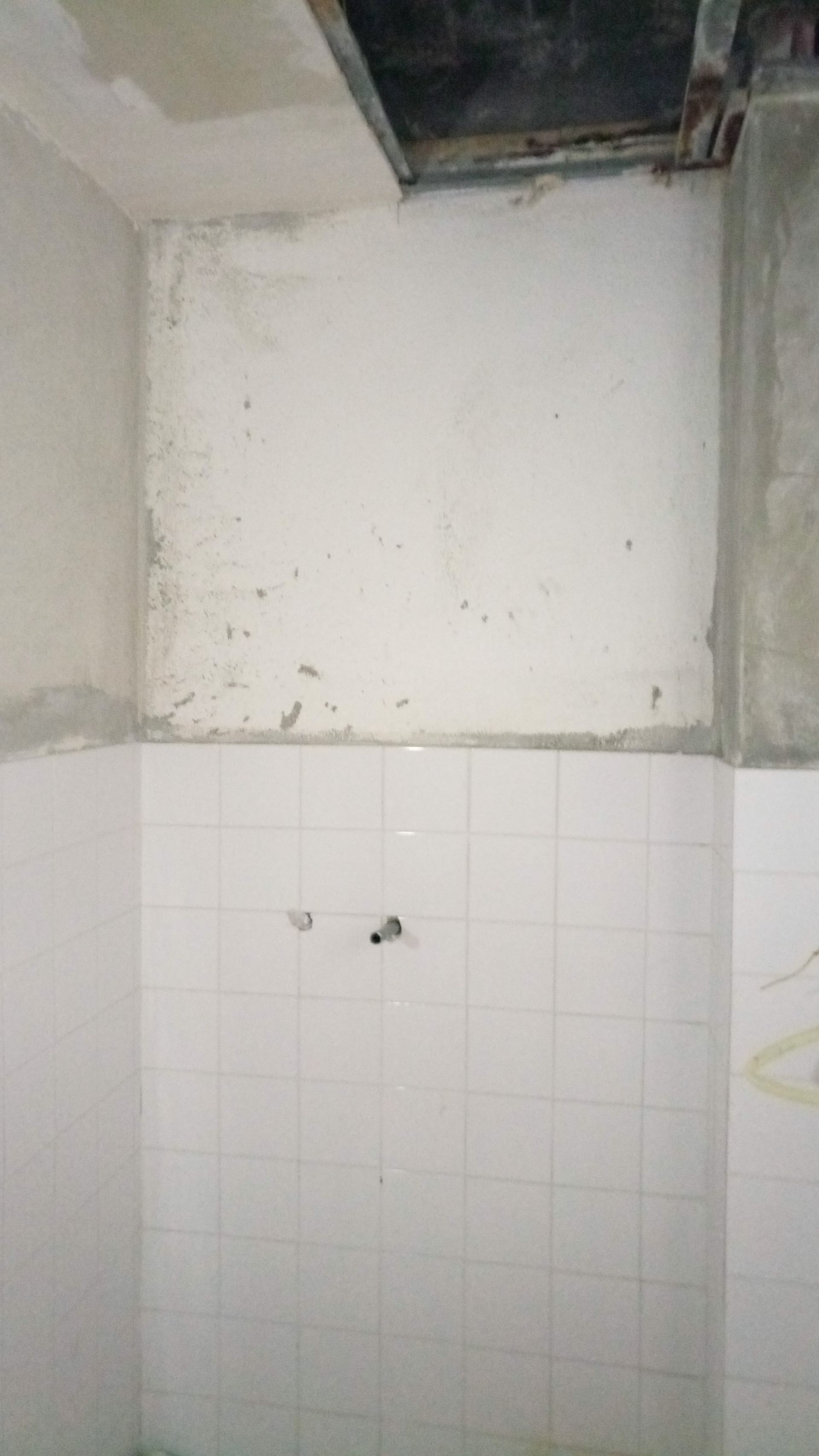 Parede revestida com azulejos brancos, com uma seção de gesso danificado e teto exposto.